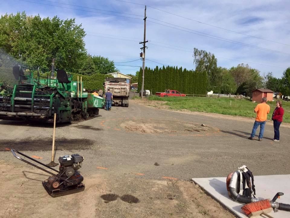 Road paving in progress: Green asphalt machine, truck, workers, and onlookers on a sunny day.