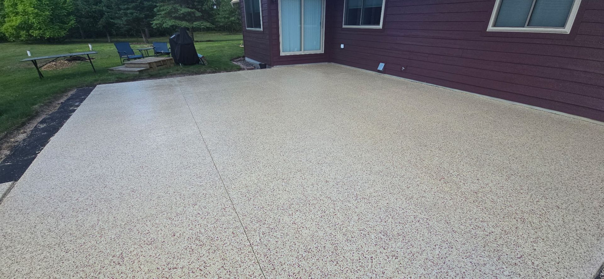 A light-colored concrete patio in front of a dark red house, with grass and a few chairs in the background.