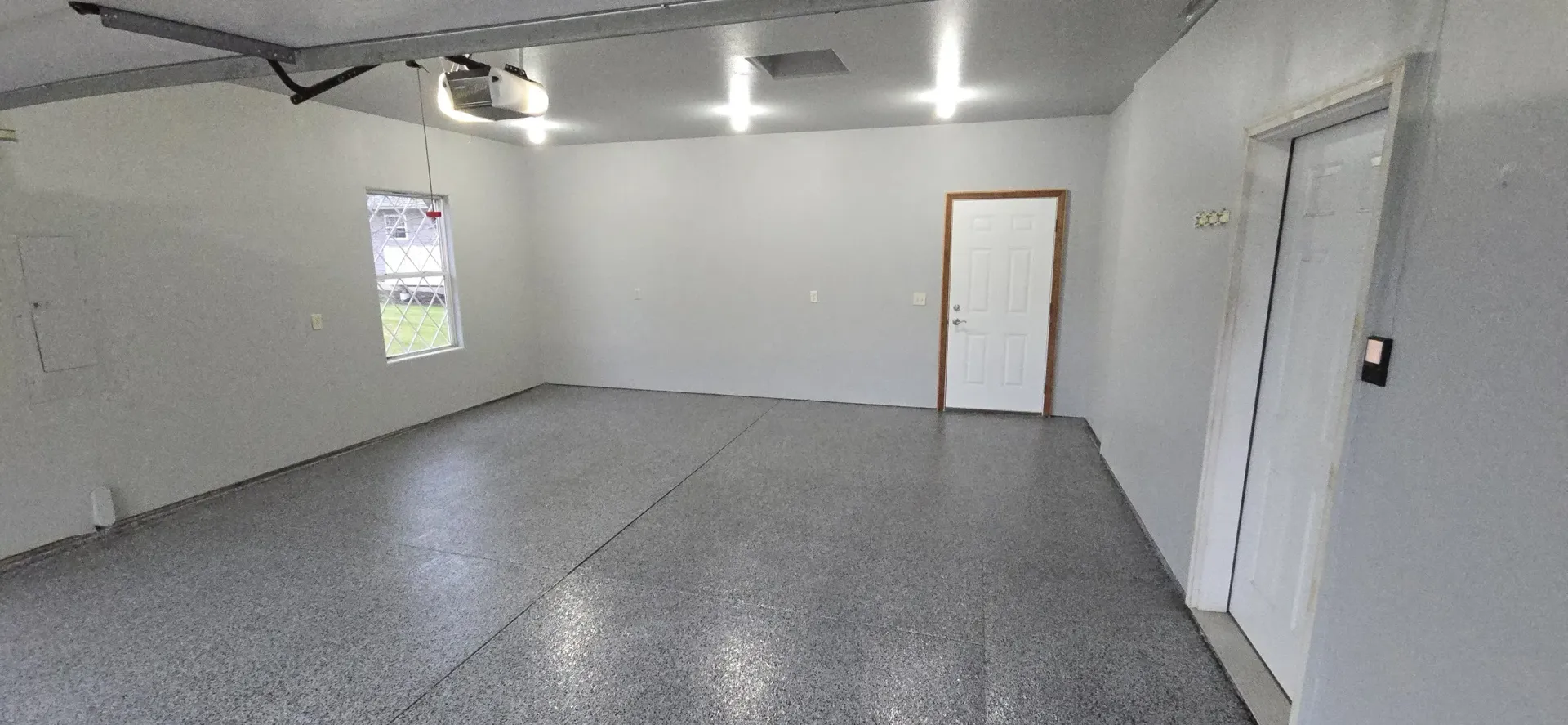A spacious, empty garage with a speckled grey floor, white walls, and a window. A white door is visible.