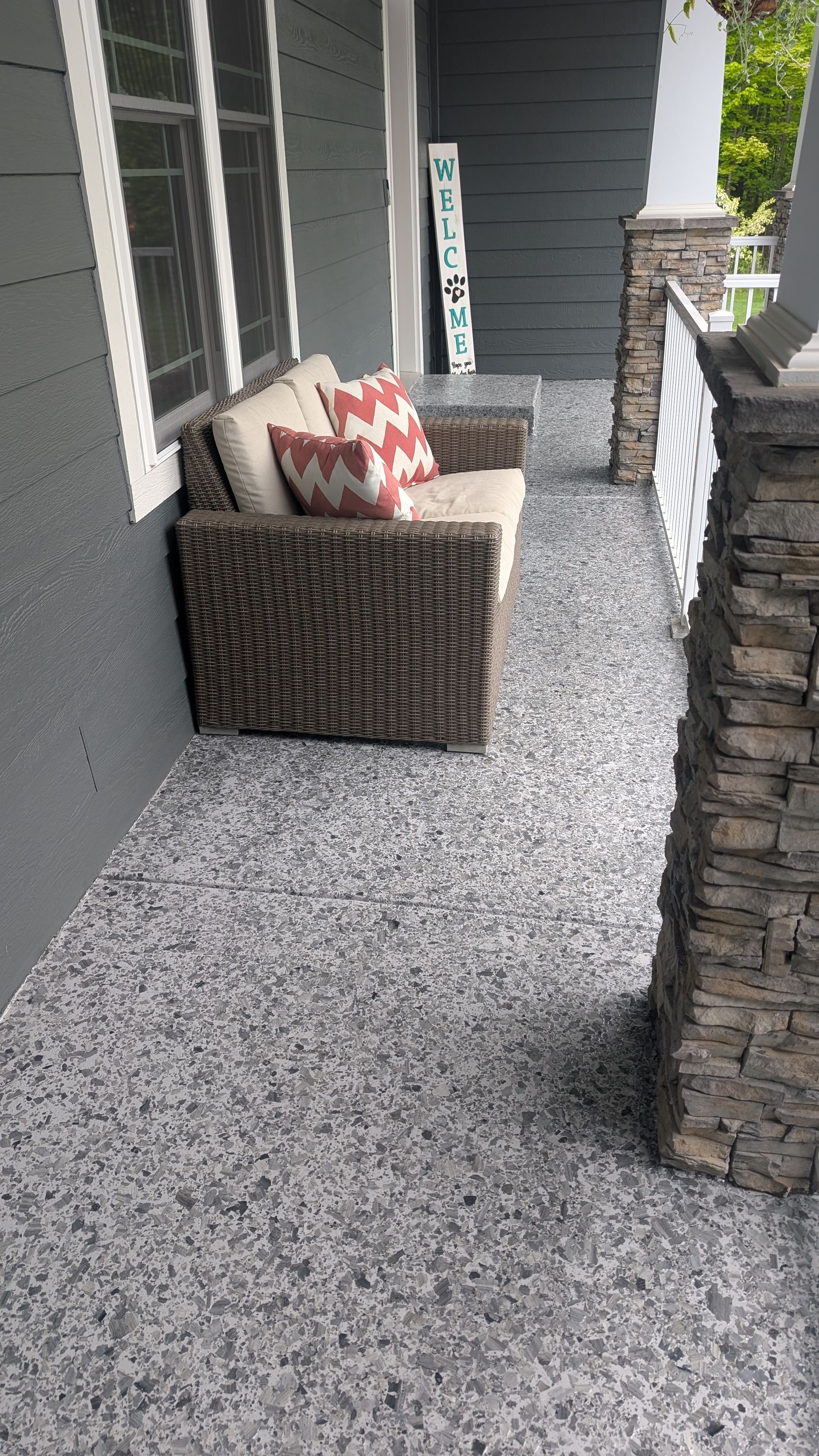 Wicker sofa with pillows on a front porch with stone columns and a gray speckled floor.