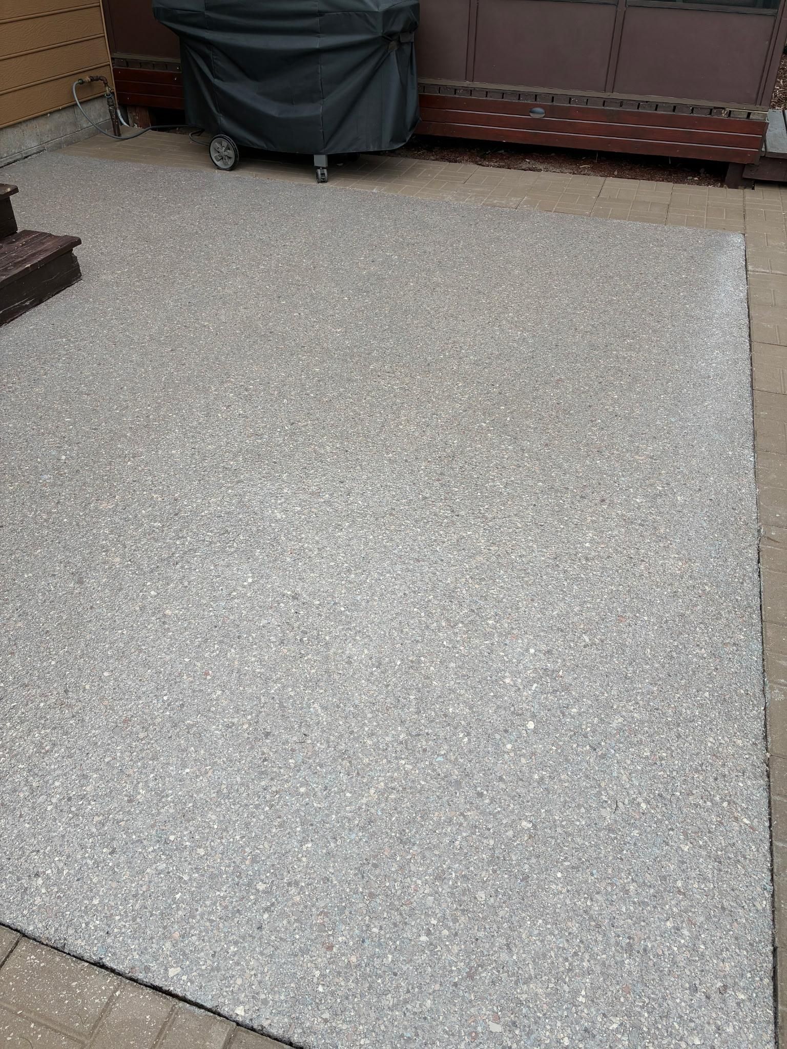 Gravel patio with a covered grill, next to a wooden deck, in an outdoor setting.