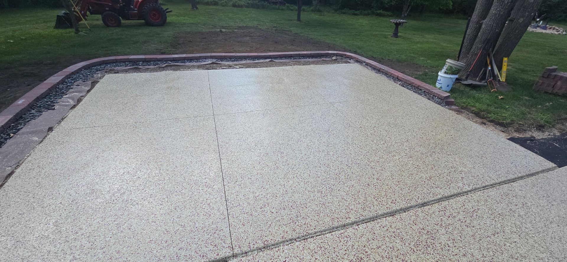 An outdoor patio made of small light-colored stones with a brick border. A red tractor sits in the distance.