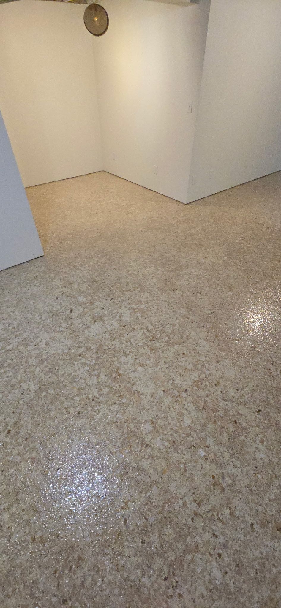 A room with a speckled floor and white walls. A circular light fixture is visible on the wall.