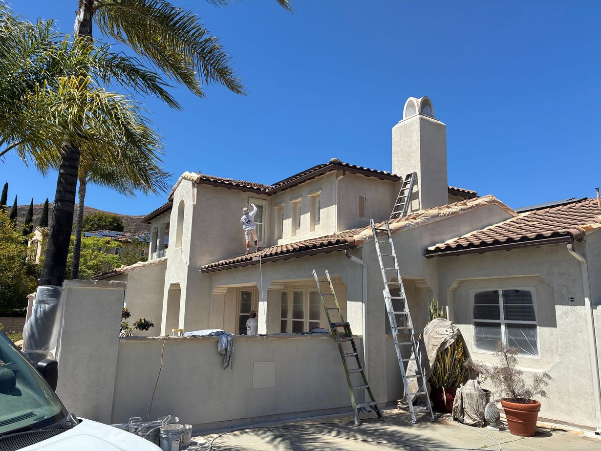Stucco house with tiled roof, ladder against wall, palm tree, and clear blue sky