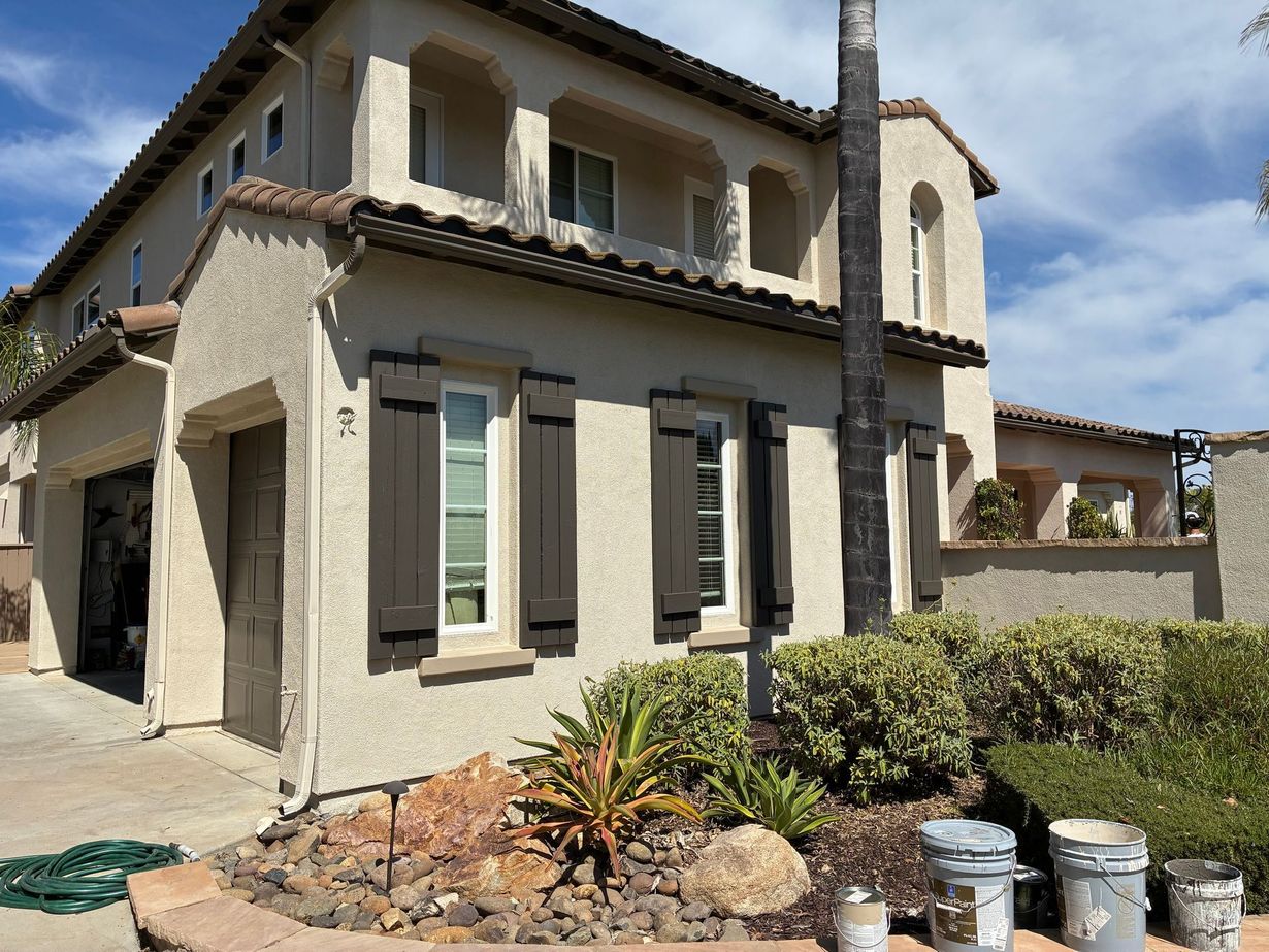 Two-story tan house with a tiled roof, covered patio, ladder, and blue recycling bins on a sunny day.