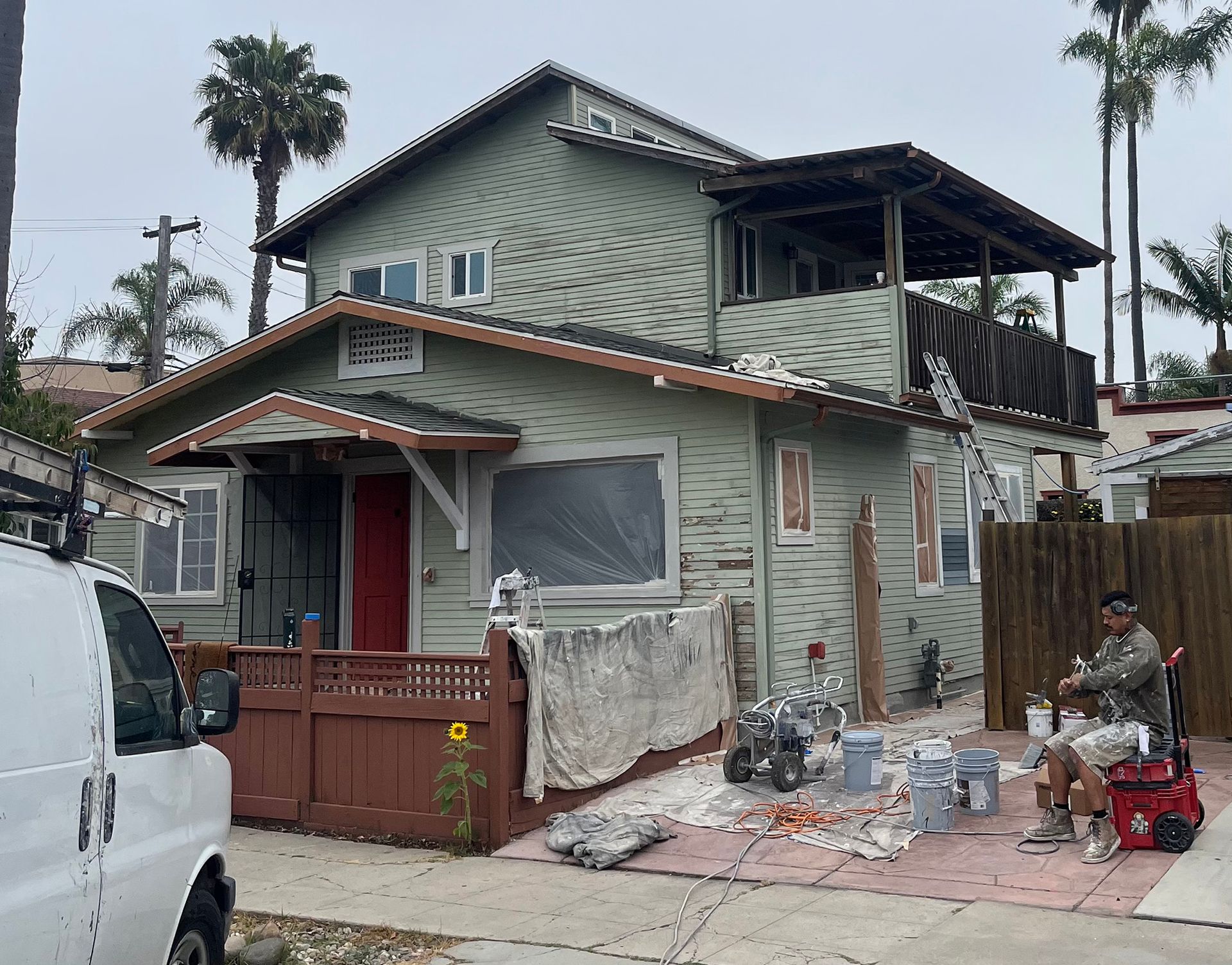 House exterior being painted, worker sitting, van parked on street.