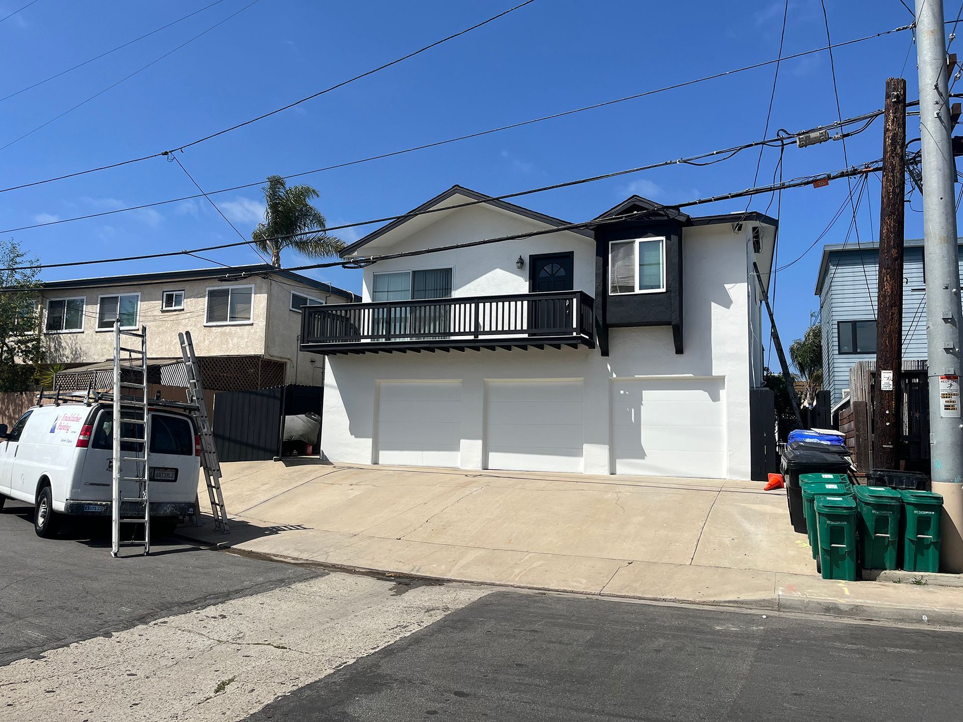 Two-story white house with balcony, black trim, and garage. Blue sky, street with parked van and trash bins.