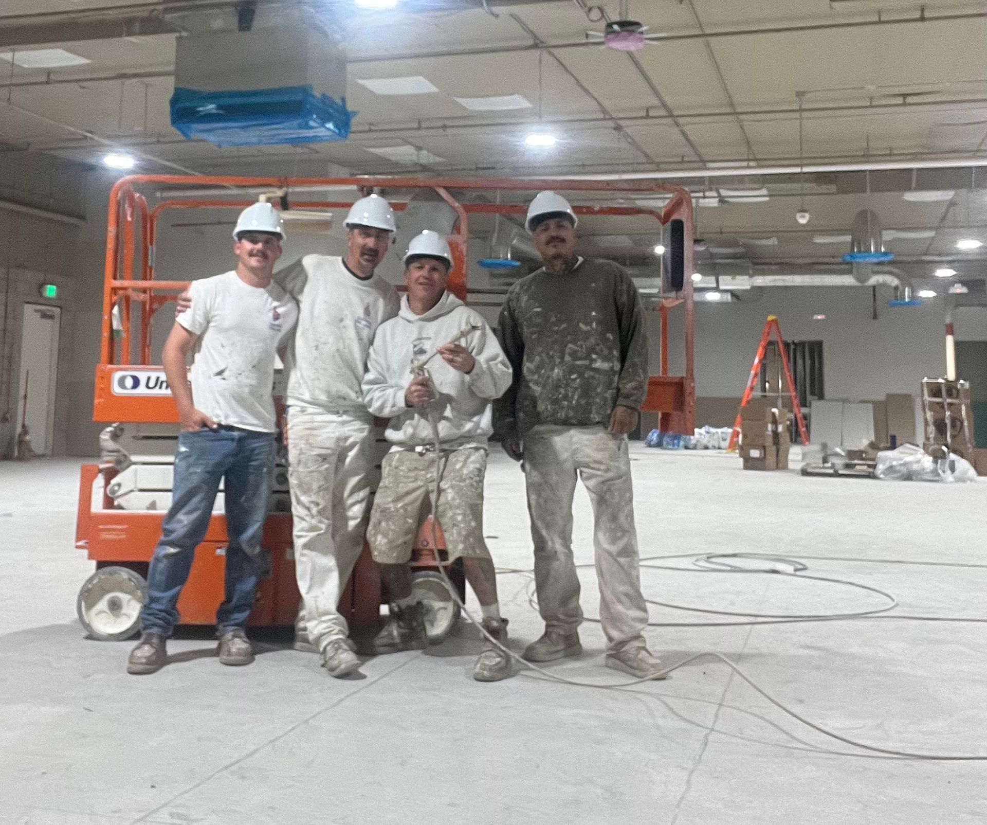 Four painters in a large, empty room pose near a scissor lift; all wear white hard hats and clothes splattered with paint.
