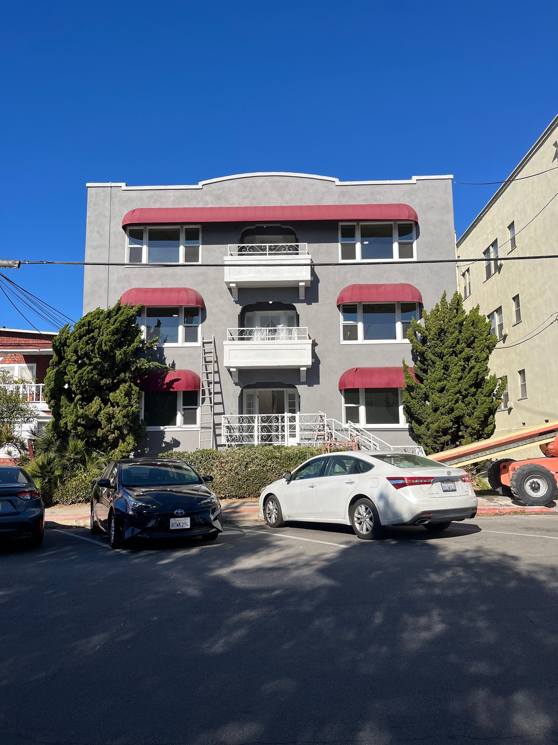 Apartment building with gray facade, red awnings over windows, cars parked in front, clear sky.