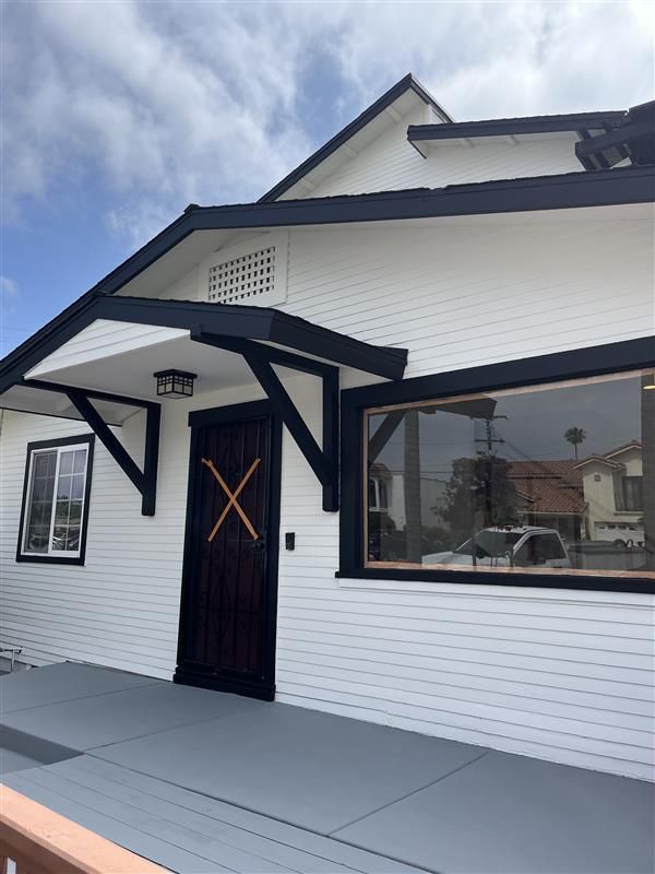 White building with black trim, a dark wood door, large window, and gray porch under a cloudy sky.