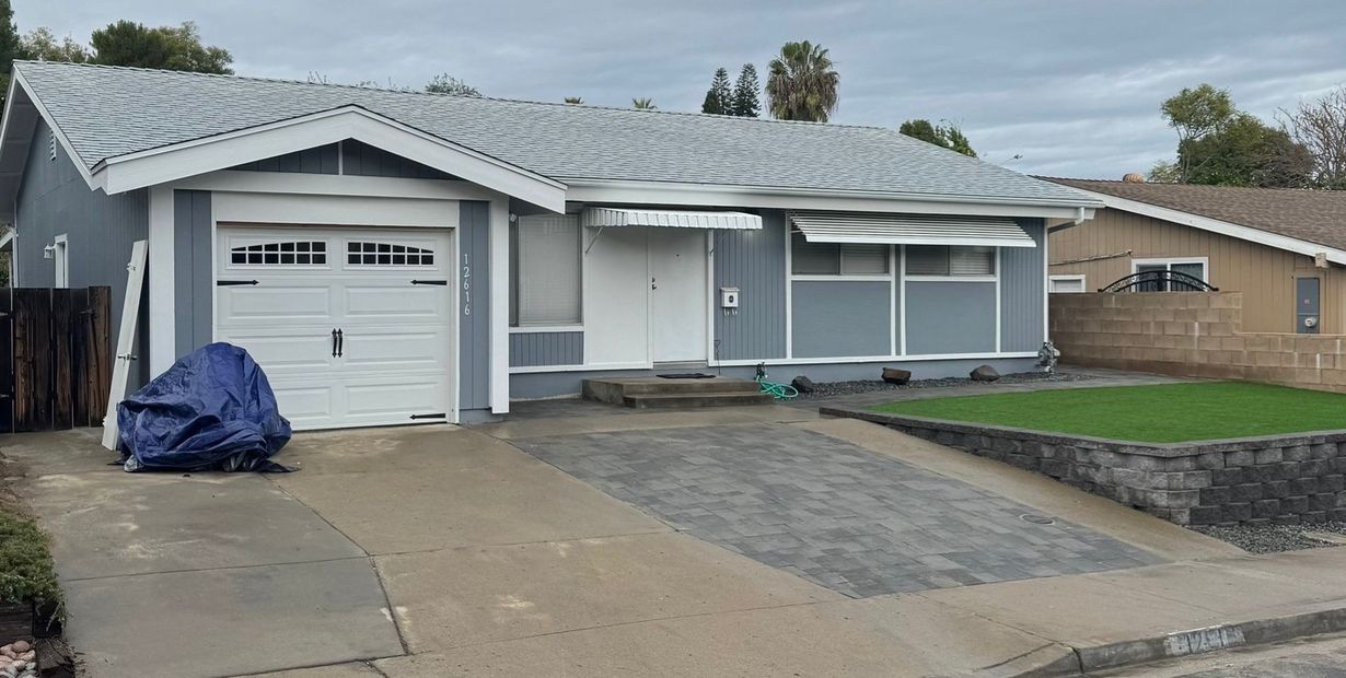 Gray house with white garage door and awning. Dark driveway, blue tarp covering something. Green grass and cloudy sky.
