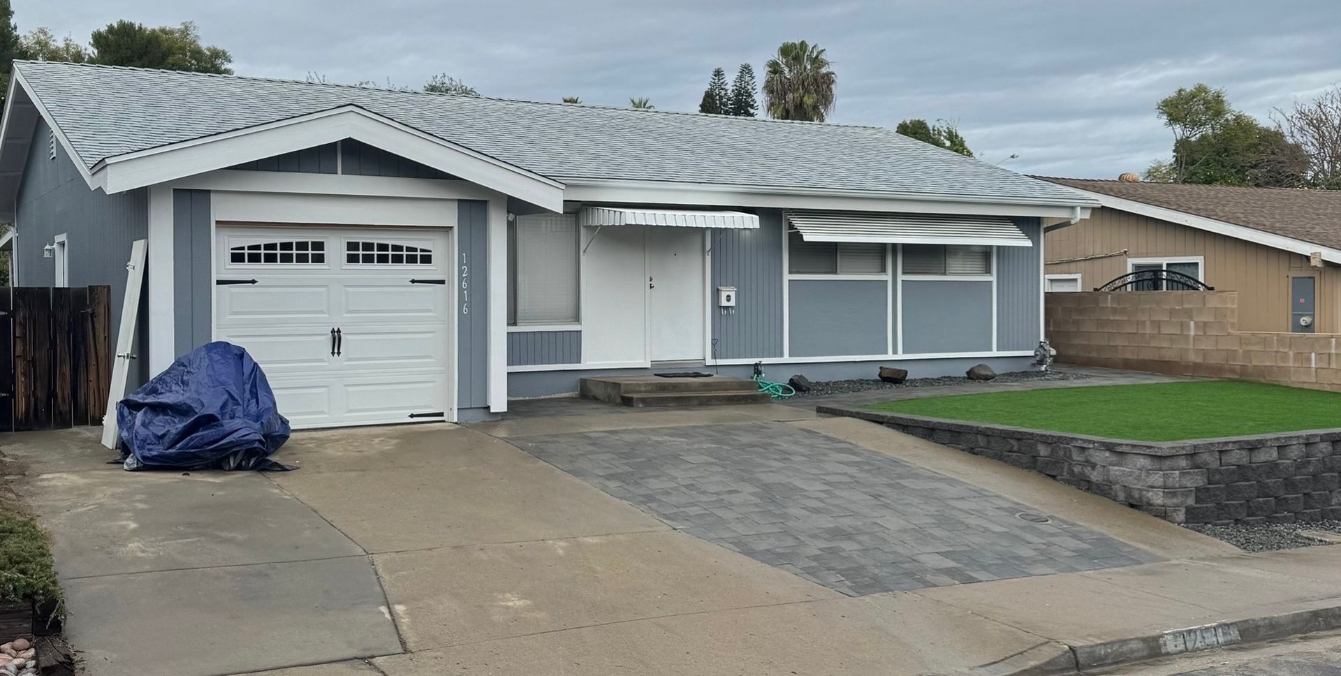 Gray house with white garage door and awning. Dark driveway, blue tarp covering something. Green grass and cloudy sky.