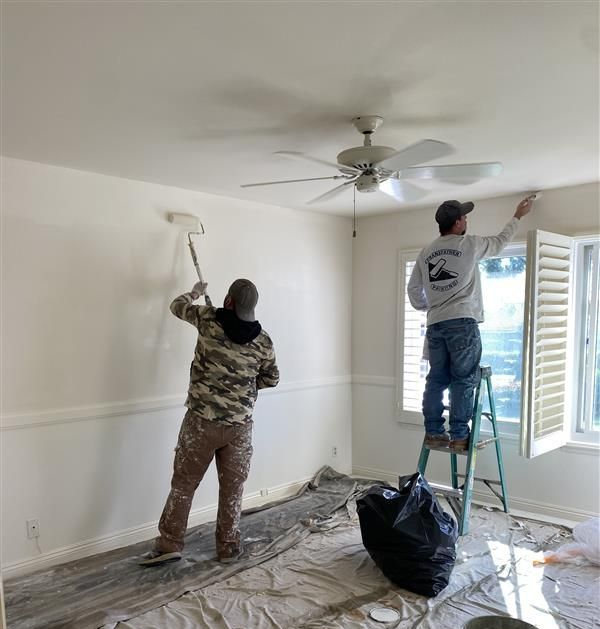 Two people painting a room one uses a roller, the other brushes near a window. Interior setting.