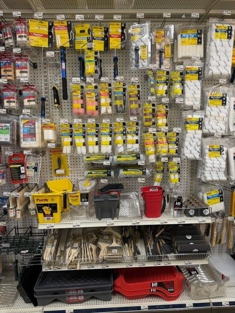 Paint supplies displayed on a store shelf, including brushes, rollers, trays, and tools, primarily yellow, black, and white.