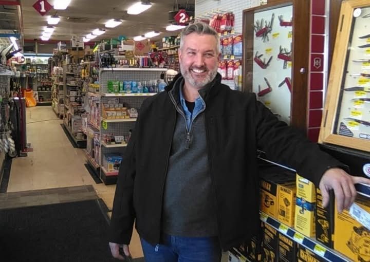 Man in a hardware store smiles, standing near shelves with tools and merchandise.