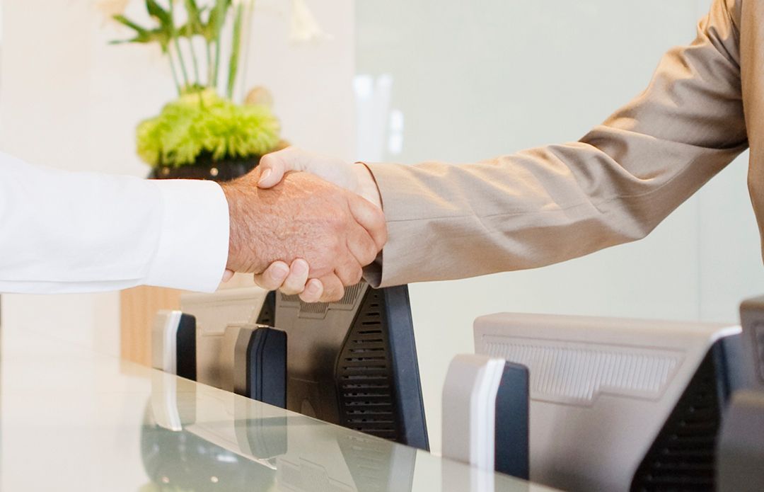 Two men are shaking hands over a desk in an office.