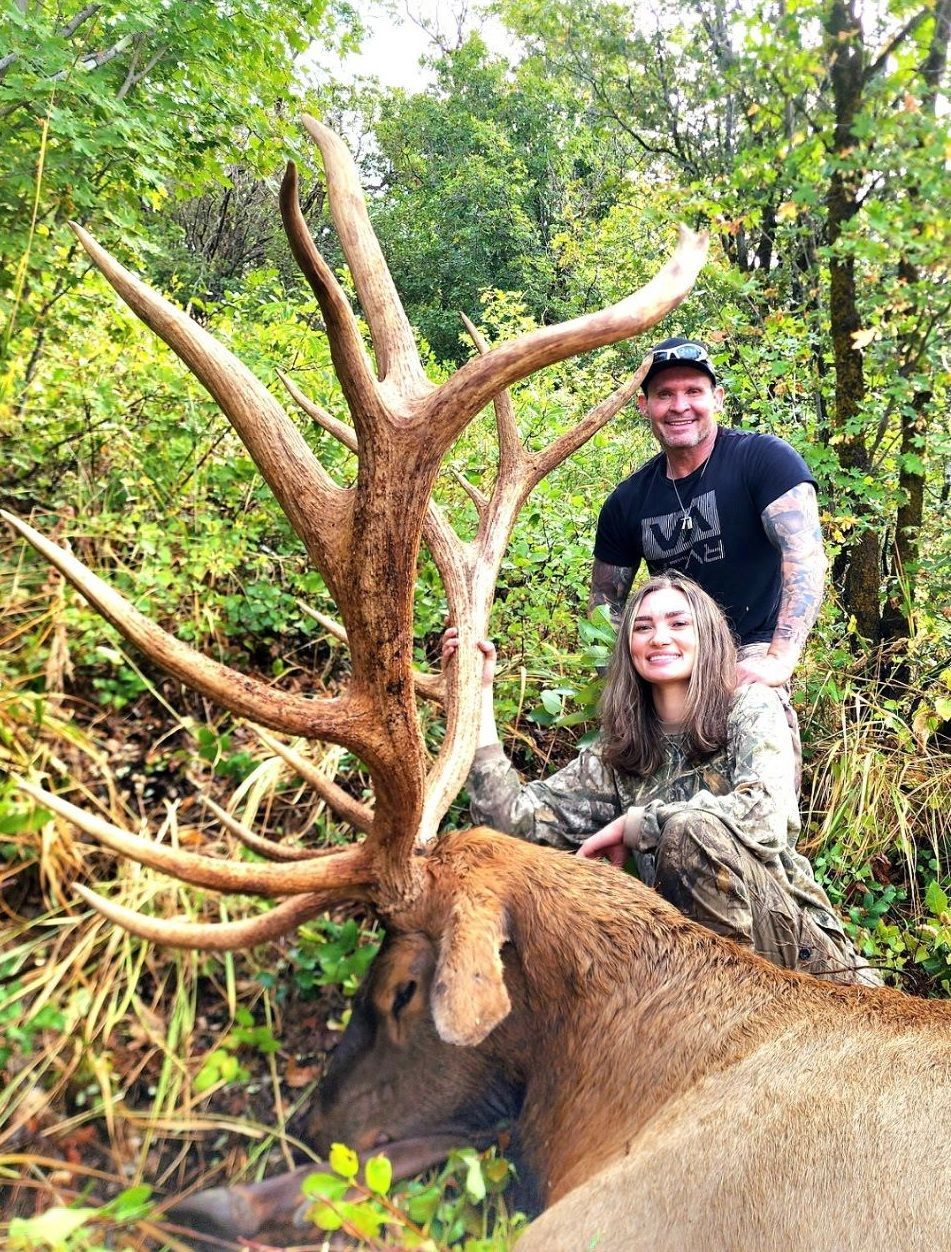 A man and woman pose with a large elk. The woman is kneeling, smiling. Green foliage surrounds them.