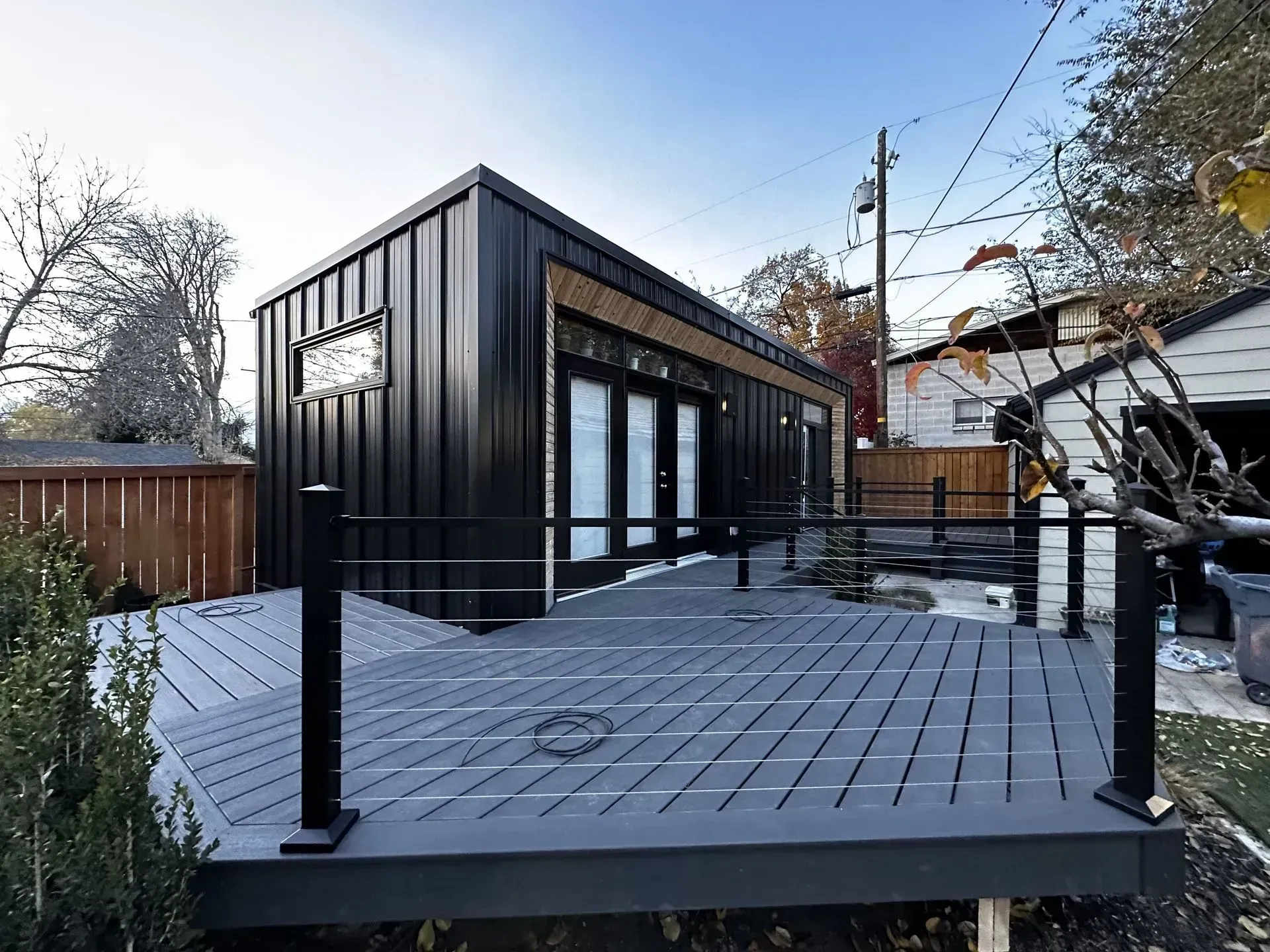 Modern black metal-sided studio building with a large grey deck and cable railing in a backyard setting.