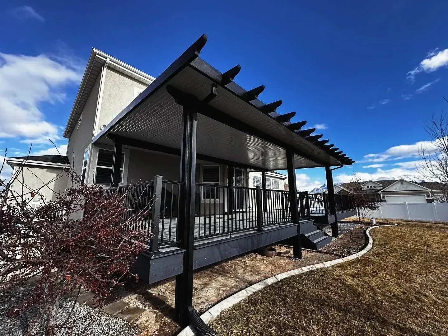 A modern home with a large, black-framed deck pergola featuring a covered patio and railings against a bright blue sky.