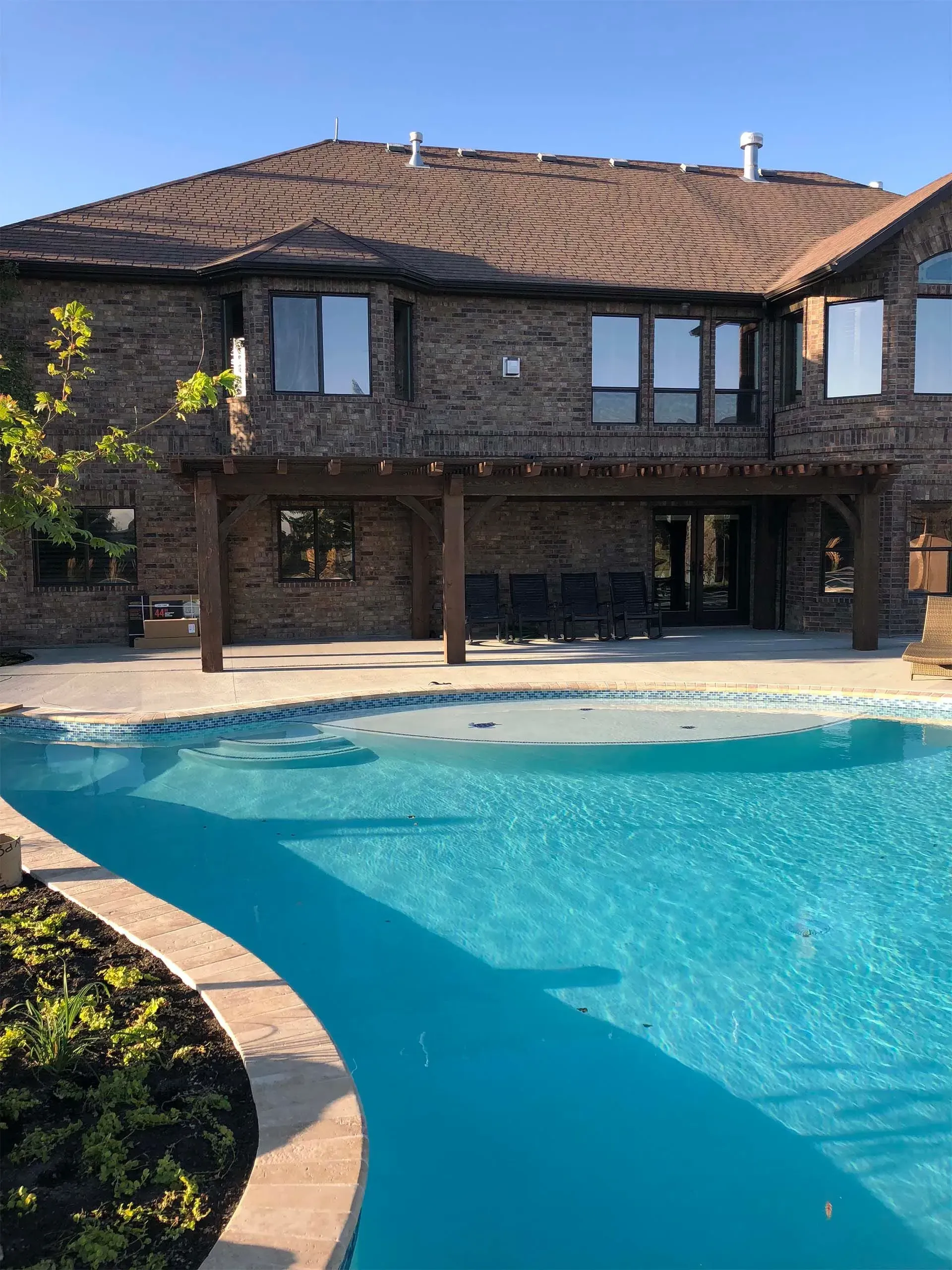 Backyard with pool, brick house, brown roof, pergola, blue water, and sunny sky.