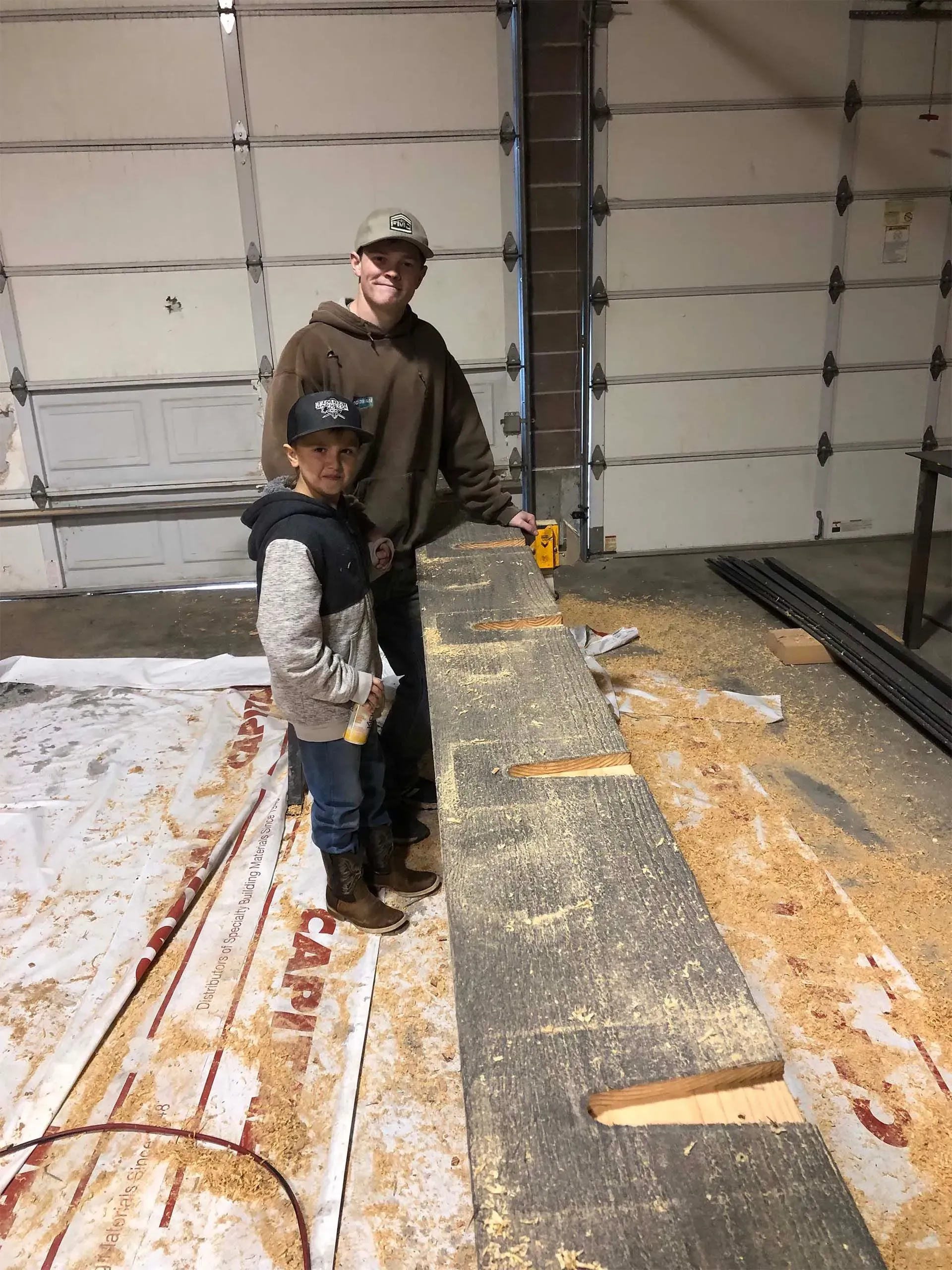Two boys woodworking in a garage. One drills a board. Sawdust covers the floor and board.