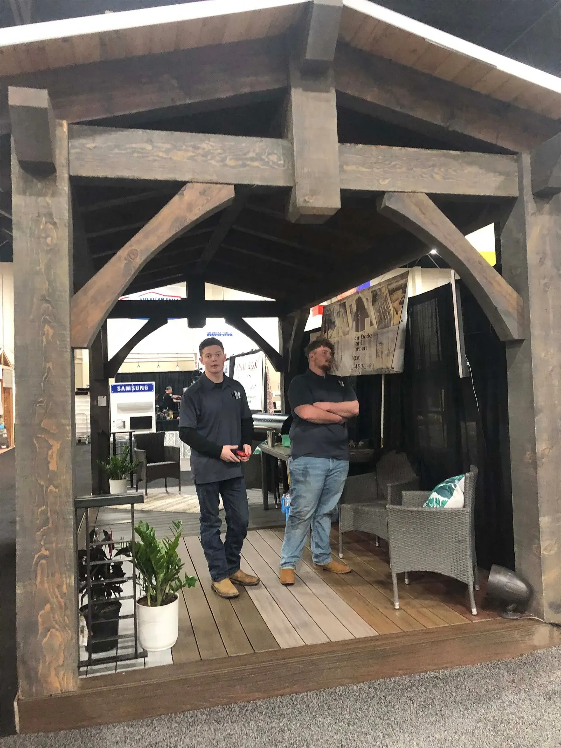 Two men stand inside a wooden structure at a trade show, looking at the camera.