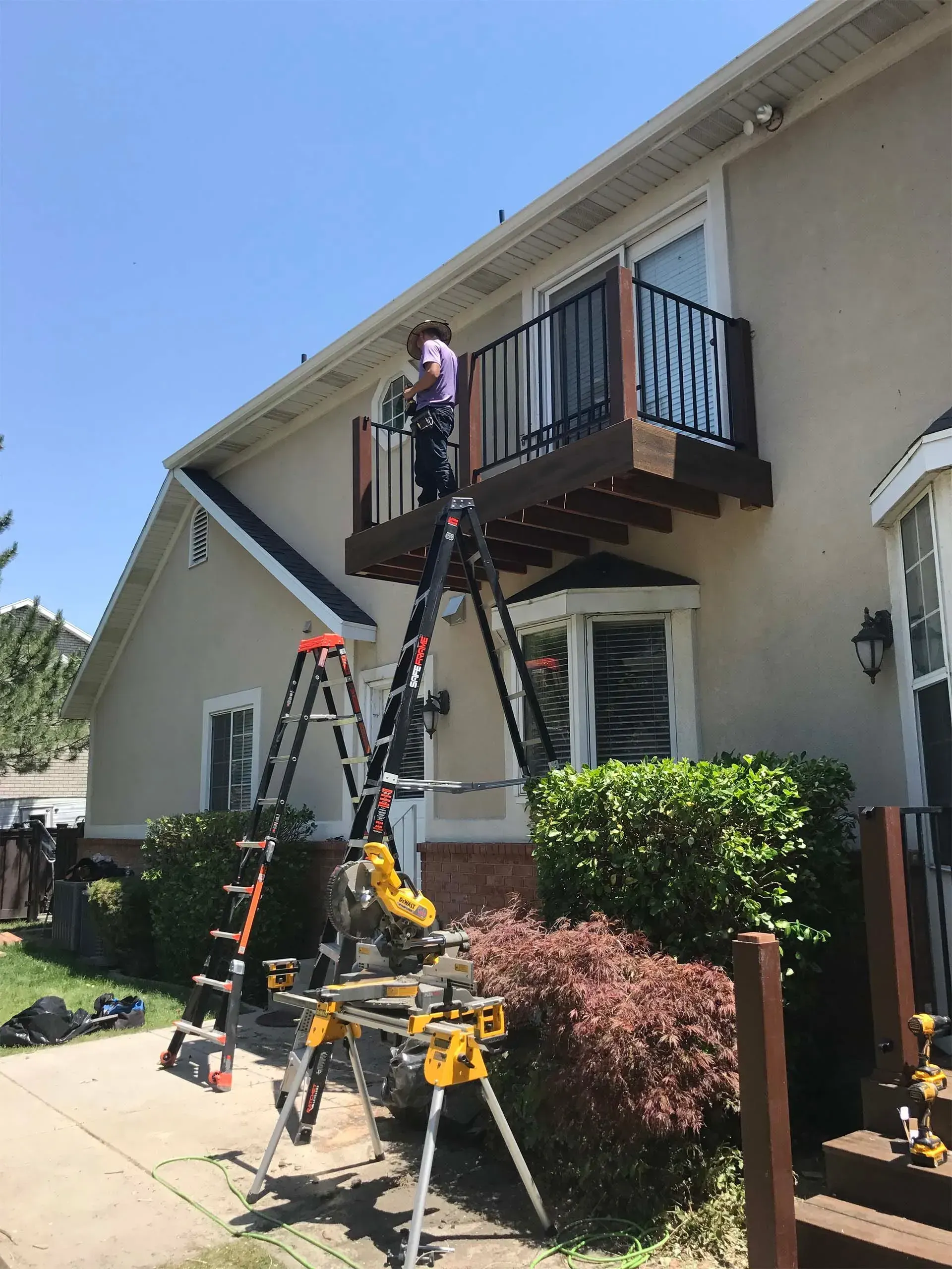 Person on a ladder working on a balcony. House exterior, sunny day. Tools and a saw horse are present.