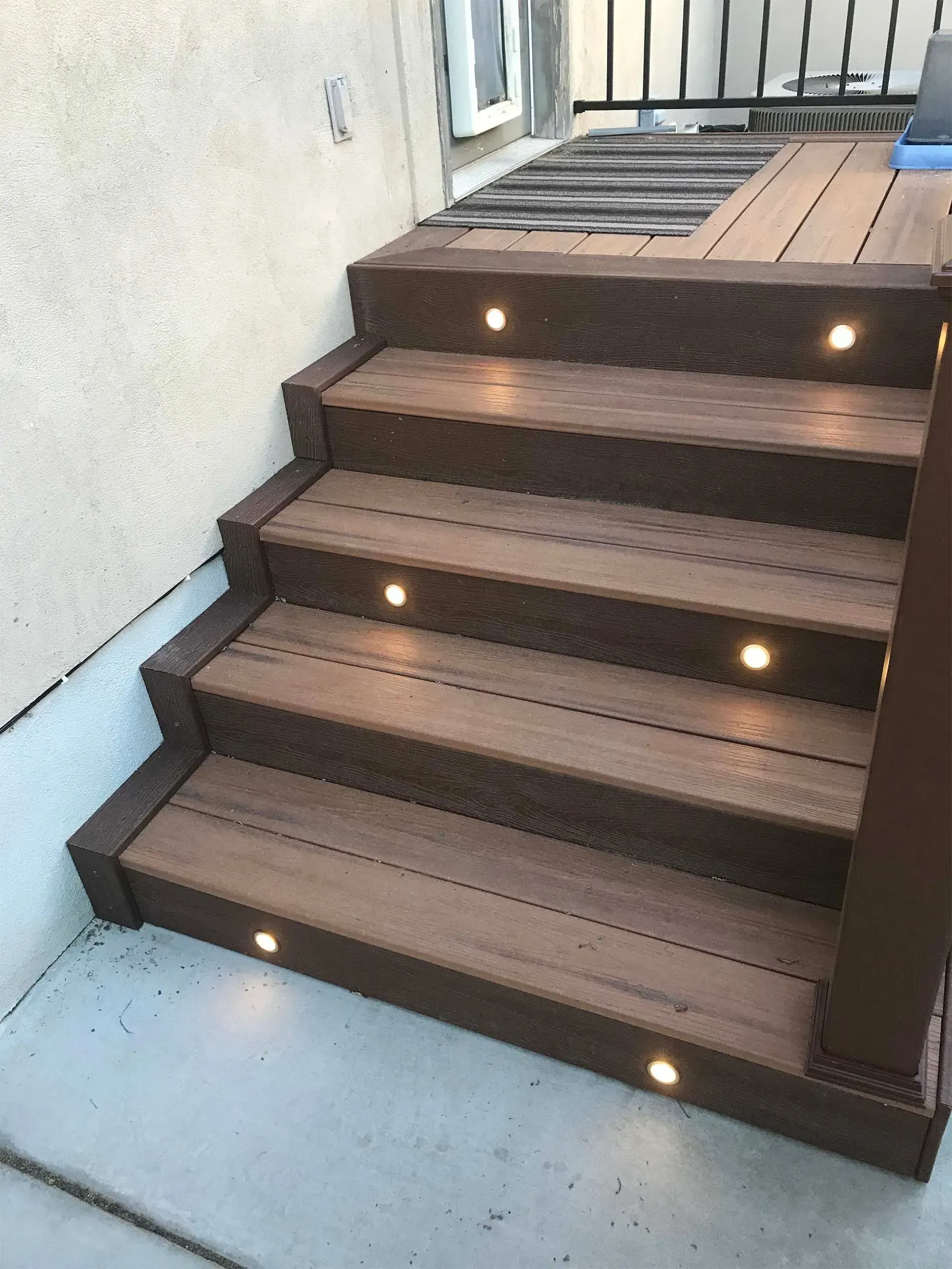 Wooden deck stairs with built-in lights, leading to a deck; against a beige wall.
