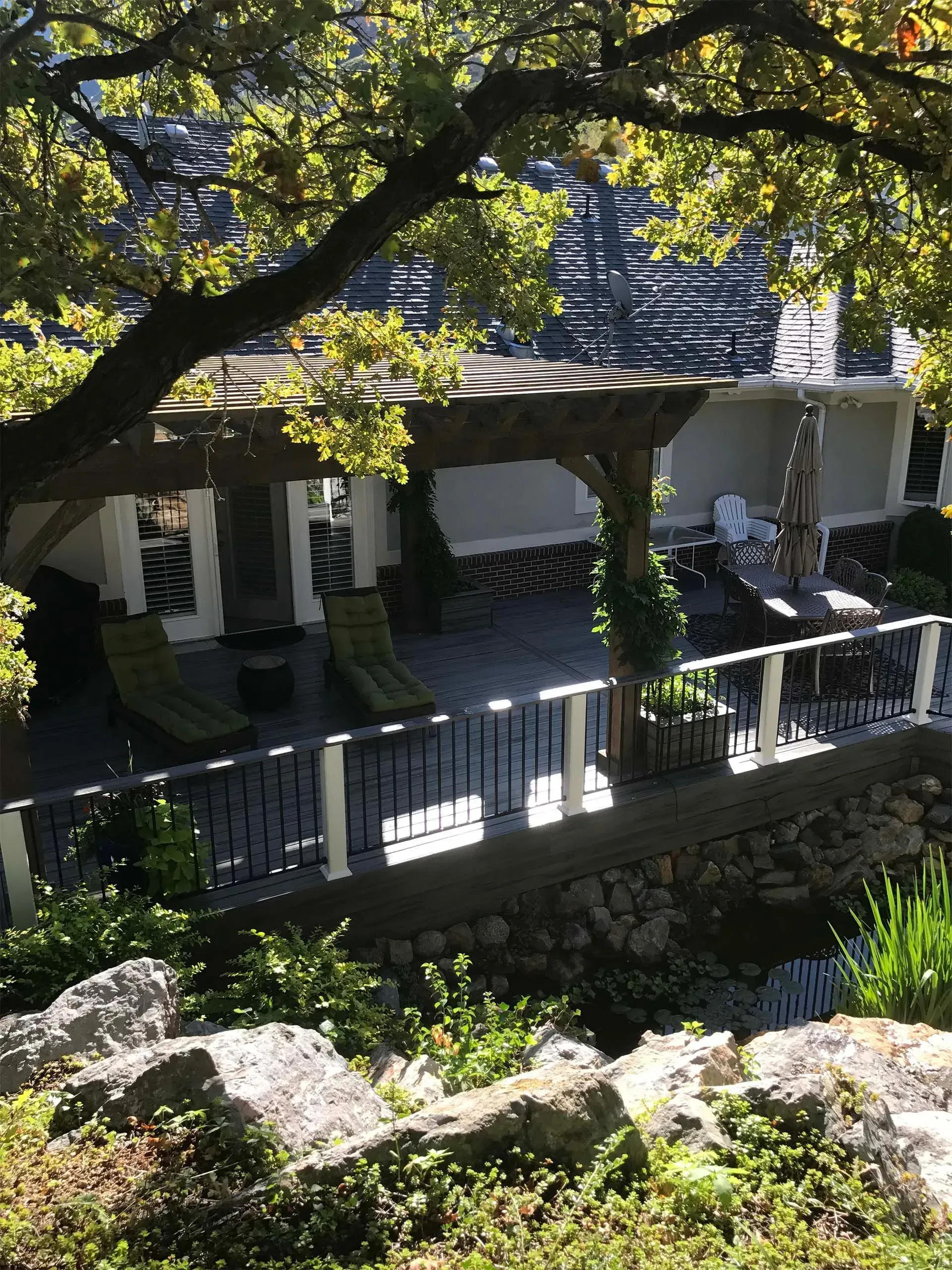 Patio with lounge chairs, pergola, and railing under a tree, with rocks and foliage in the foreground.