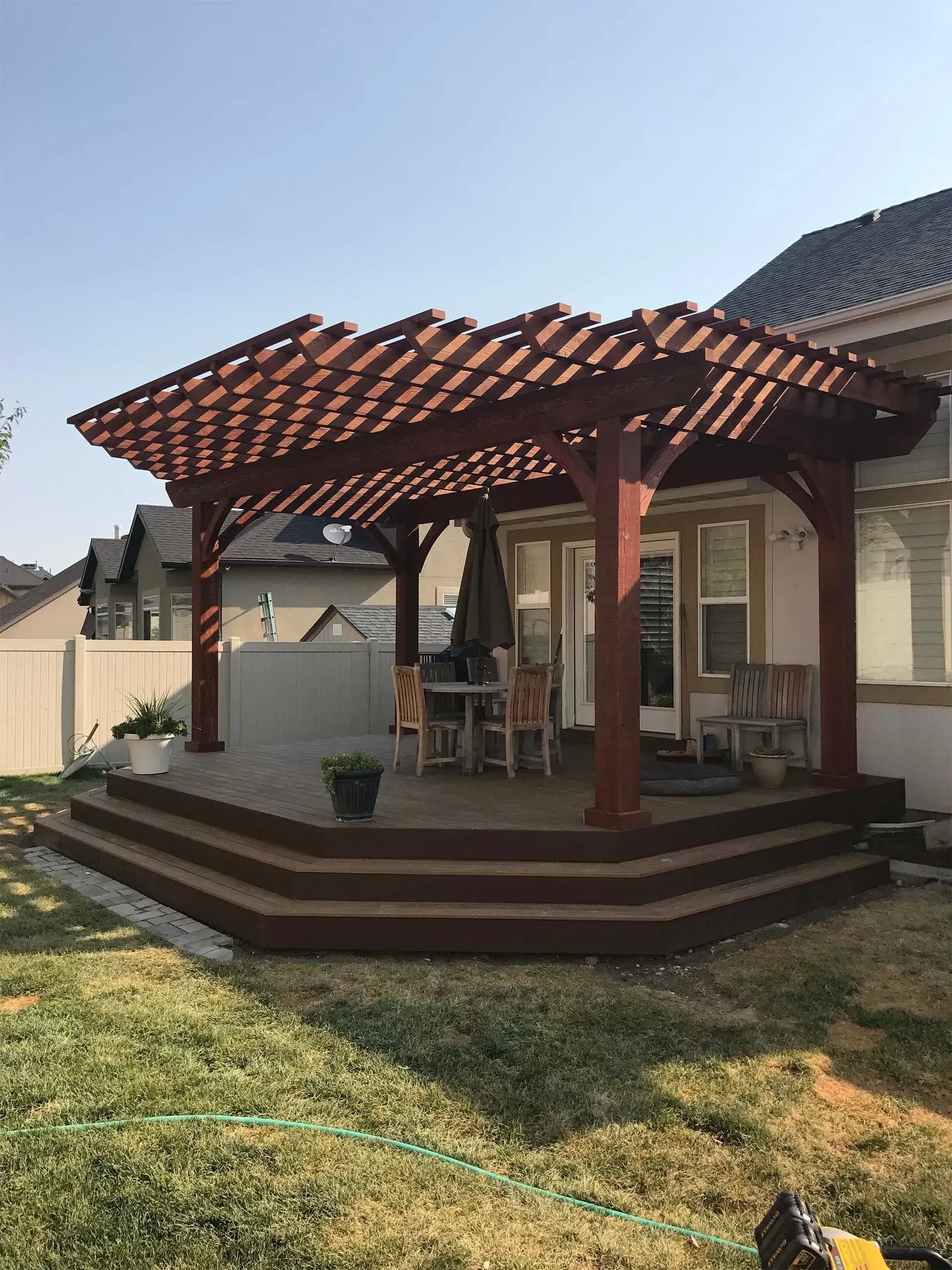 Wooden pergola over a multi-level deck with dining table and chairs, backyard setting.