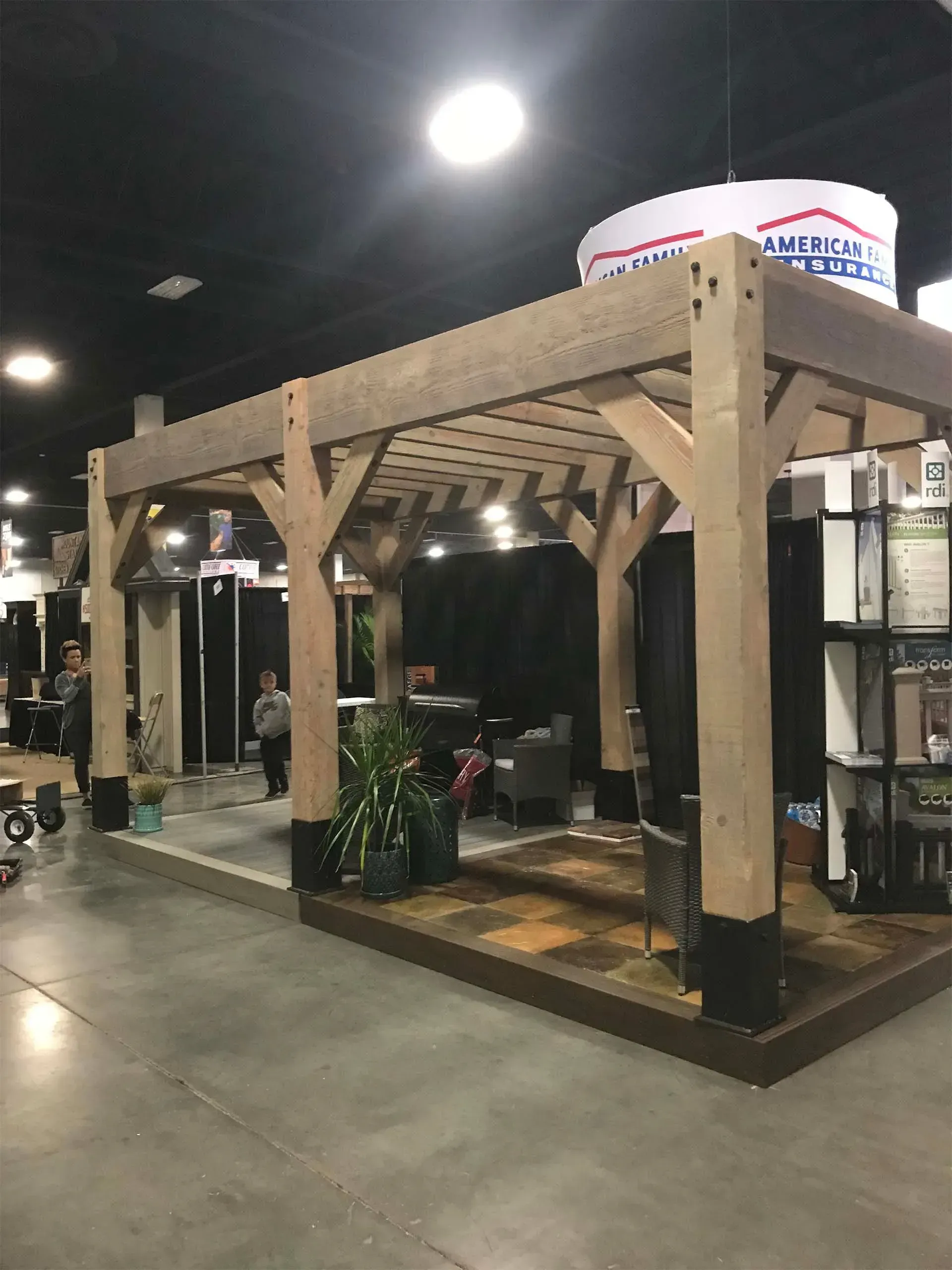 Wooden pergola structure at a trade show, set on a gray concrete floor, with a plant and dark backdrop.