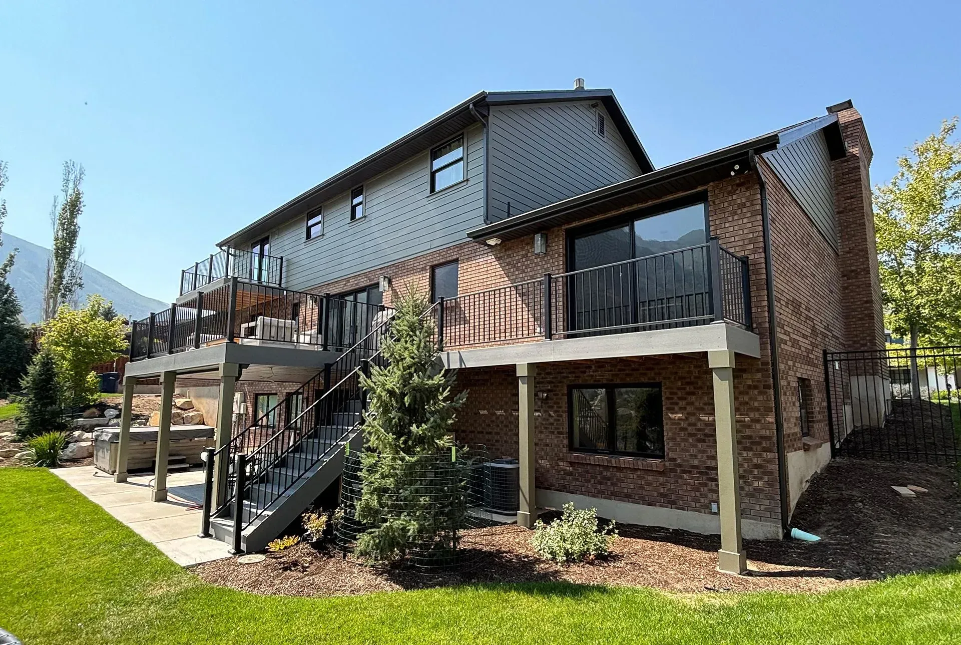 Back of a two-story house with a deck, brick facade, and dark siding. Green lawn, blue sky.