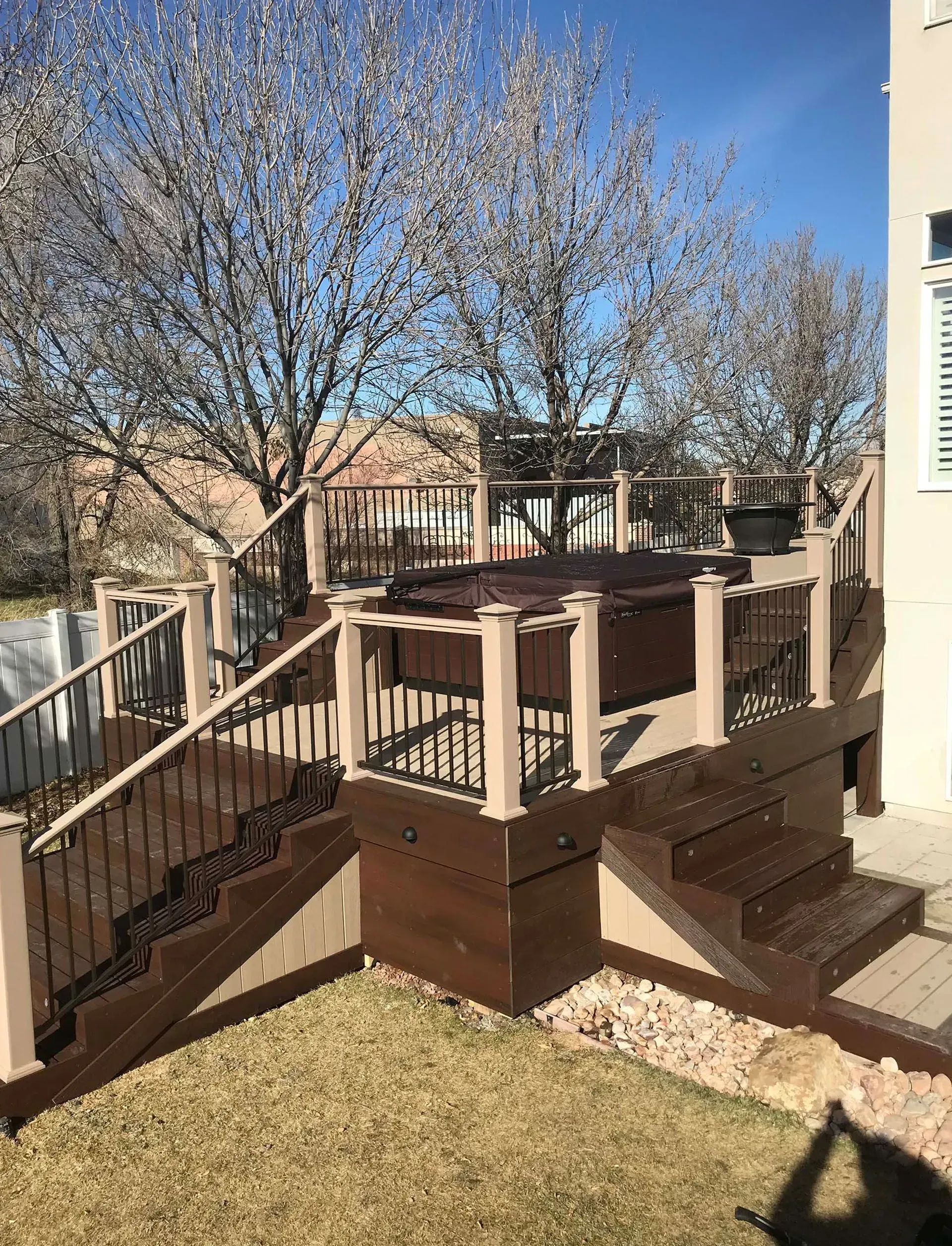 Multi-level brown deck with hot tub, railings, steps, and bare tree branches in a yard.