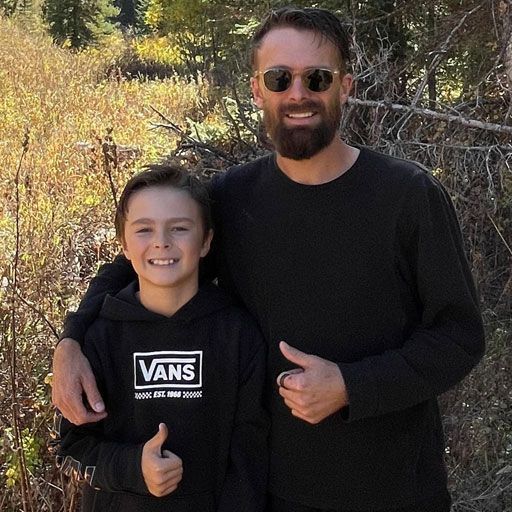 A man and a boy smiling with thumbs up in front of trees and tall grass.