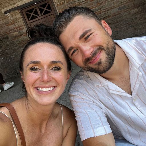 A woman and a man smiling close-up. Woman with bun, brown strap, and fair skin. Man with a beard, a striped shirt, and tan skin.