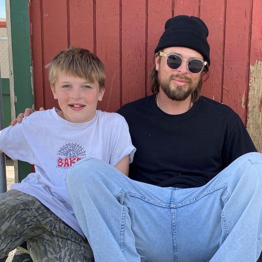 A man and a boy sit together. The man wears a black beanie, sunglasses, and black shirt, while the boy wears a white t-shirt.