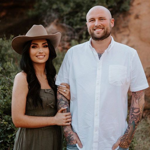 A woman in a hat and a man with tattoos pose outdoors, smiling.