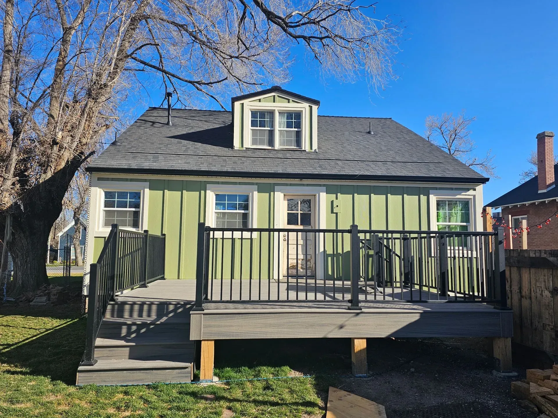 Green cottage with black railing, deck, dormer, and a tree, under a blue sky.