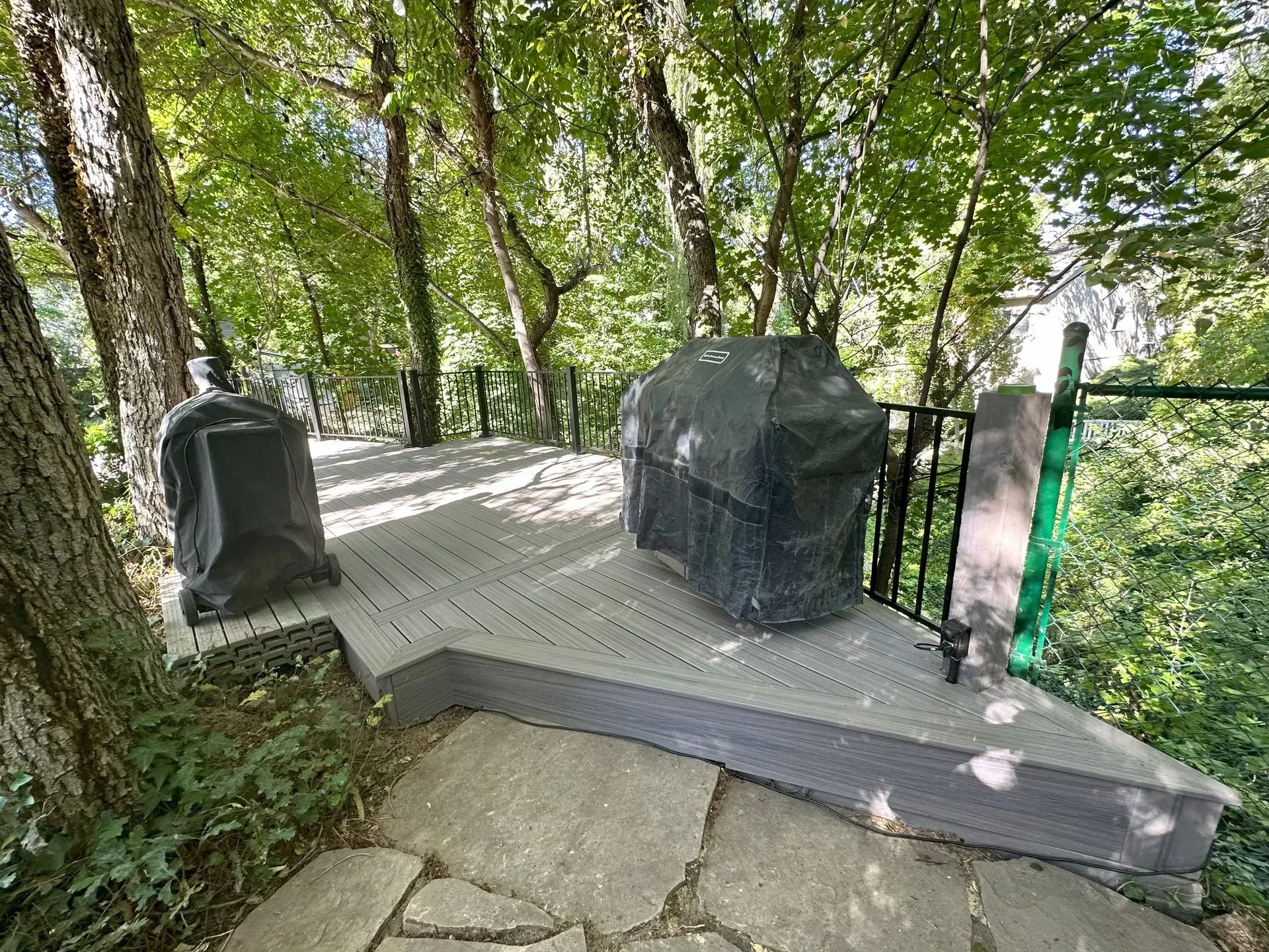 A composite deck with a covered grill and smoker, surrounded by trees, accessible via stone path.