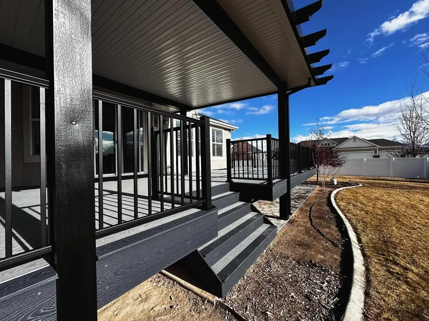 Black-railed deck with stairs, gray steps, and a covered patio overlooking a backyard on a sunny day.