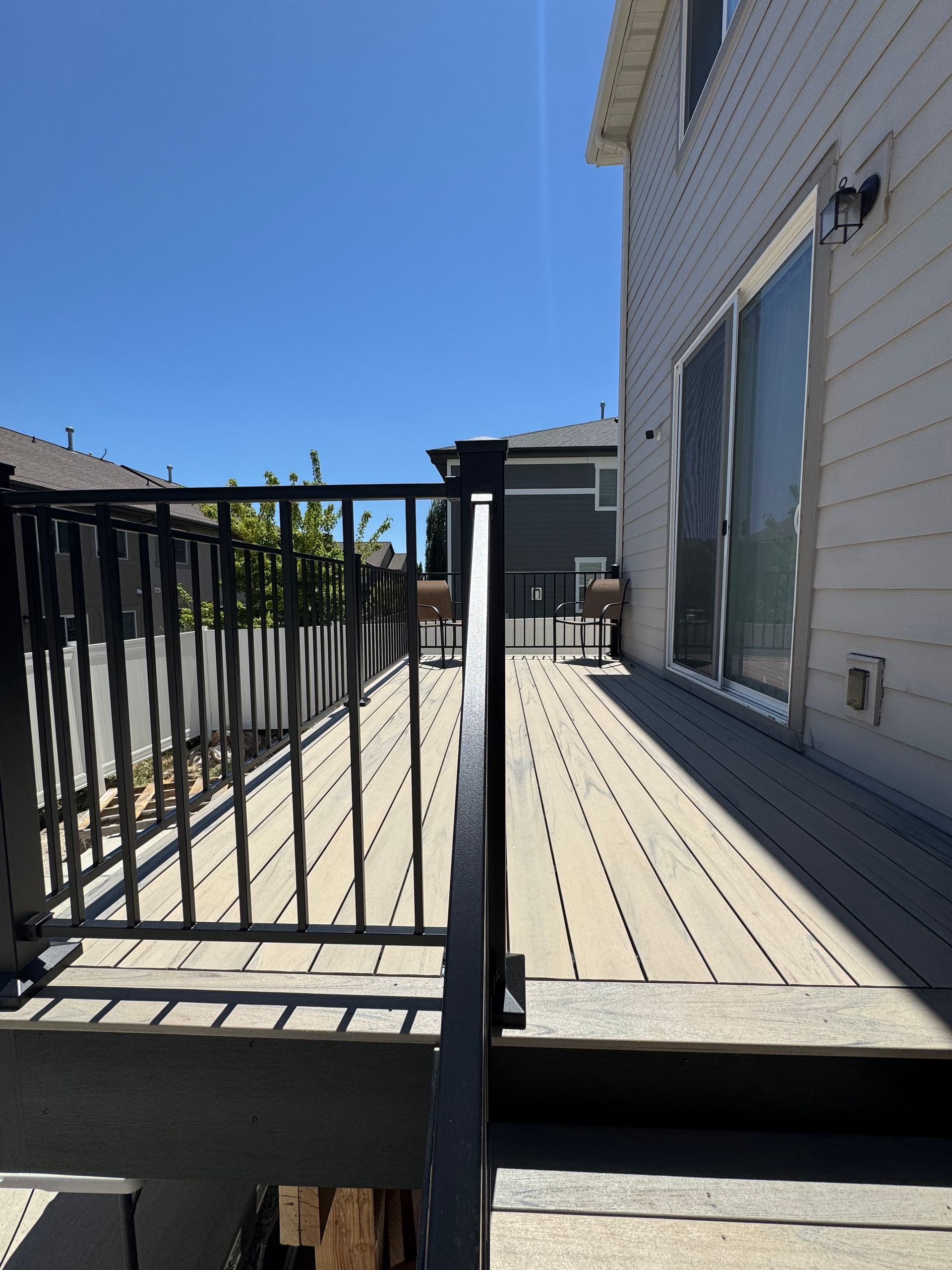 Deck with black railing, gray decking, and a sliding glass door against a sunny blue sky.