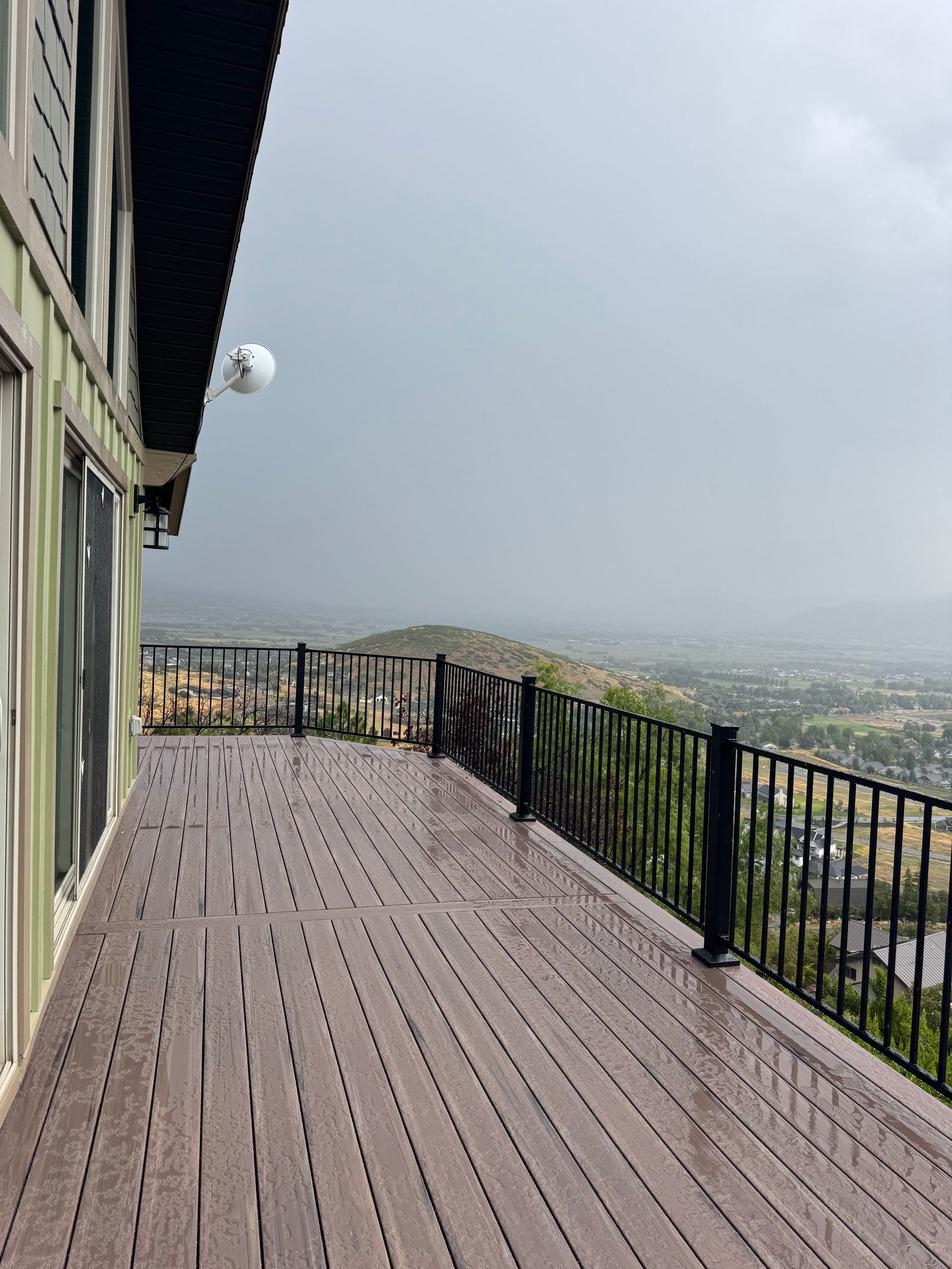 A wooden deck with a black railing overlooks a cloudy, mountainous landscape. The deck is next to a house.