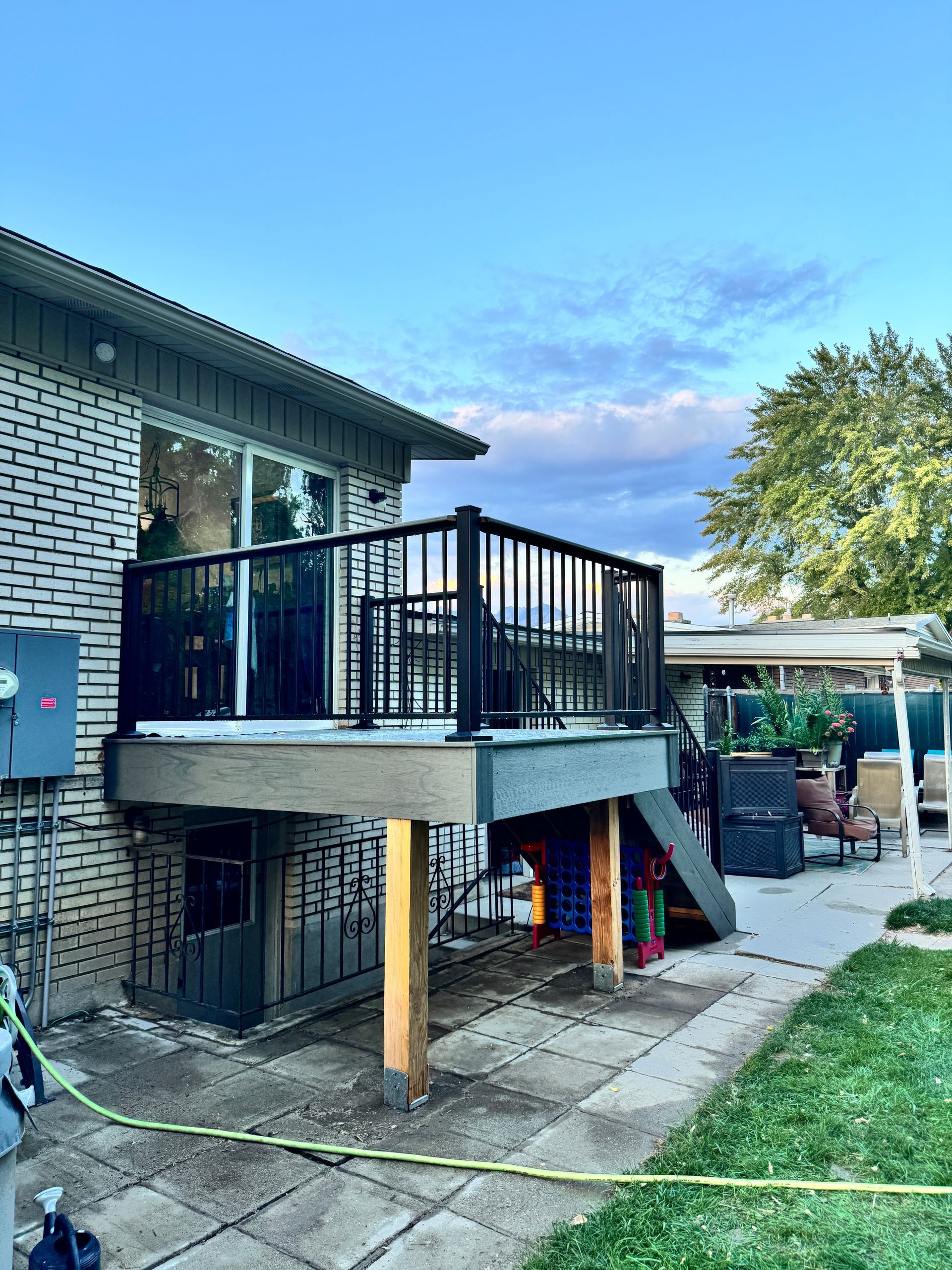 A backyard deck with black railing, attached to a brick house, stairs, and a paved patio.