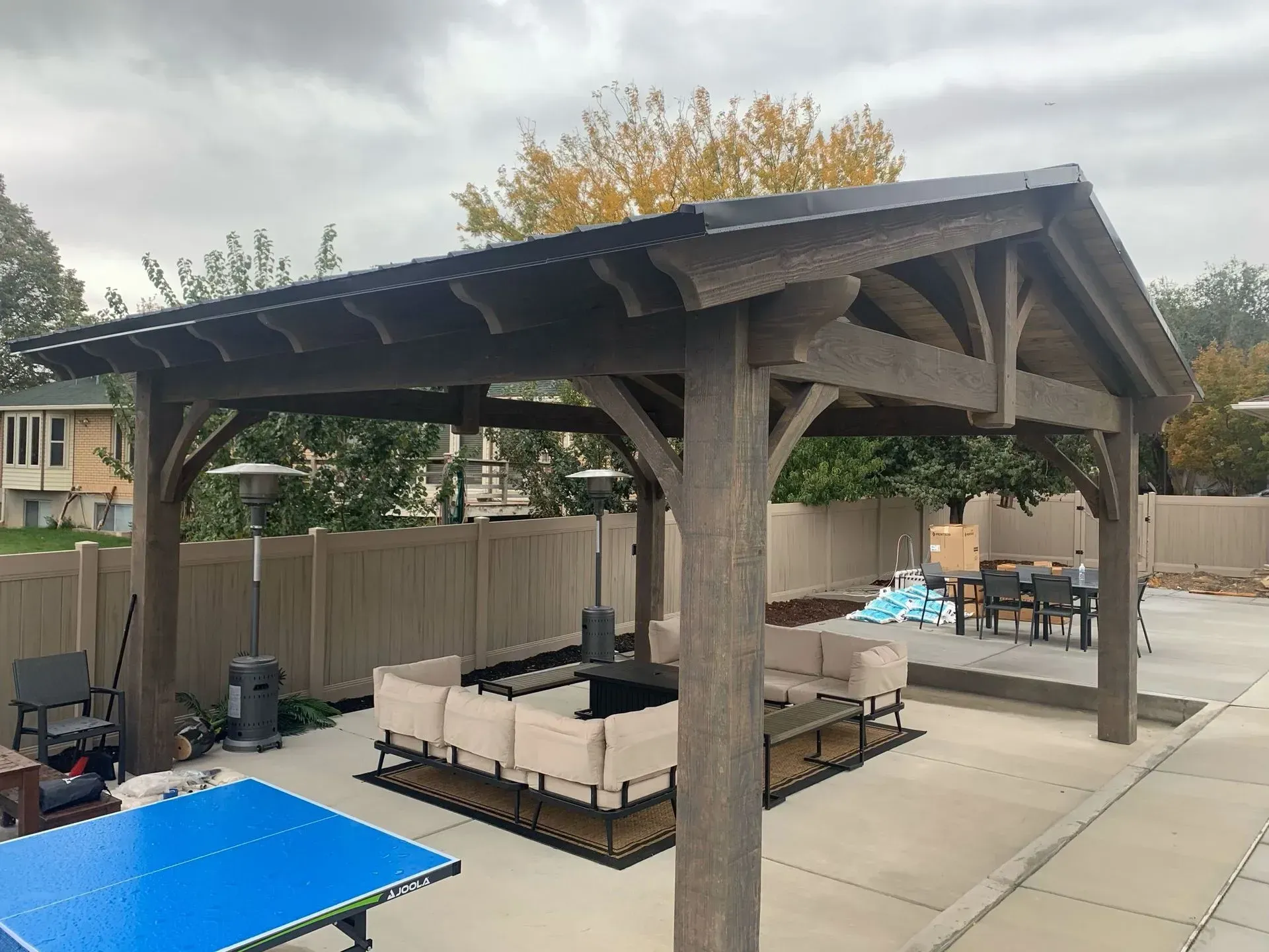 A wooden pergola with seating, fire pit, and heaters in a backyard setting.