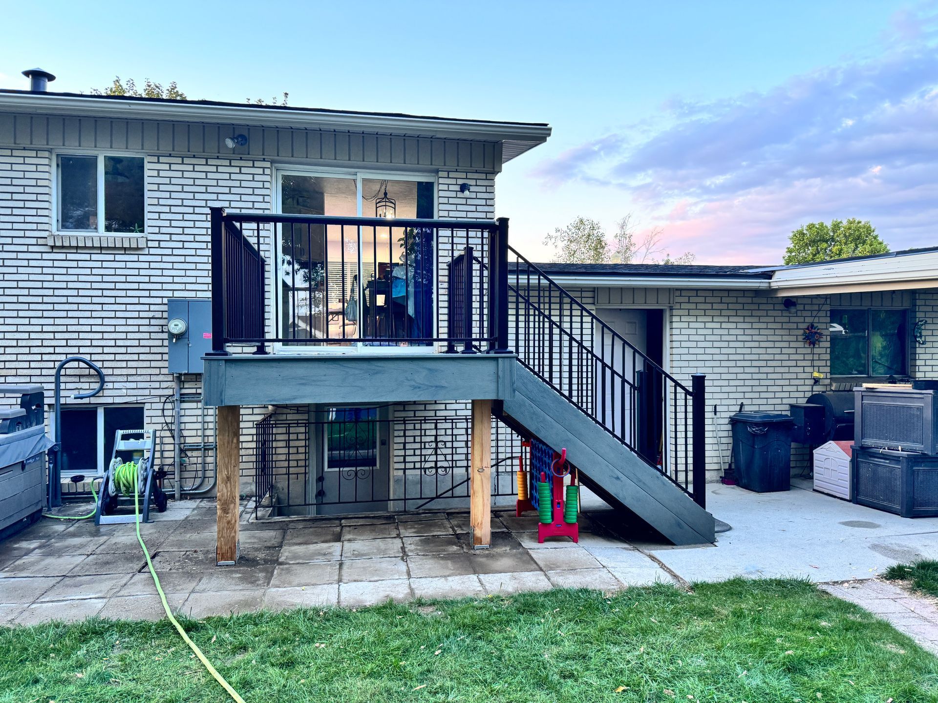 A gray deck with black railing attached to a brick house, with a staircase leading down to a patio.
