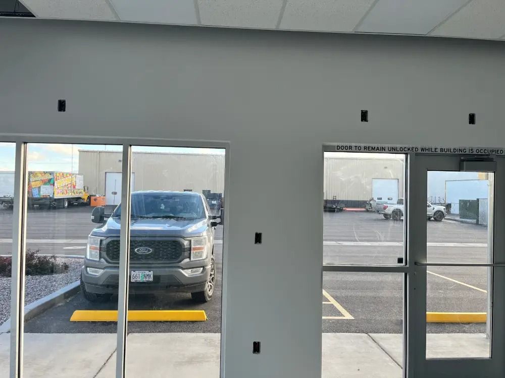 A gray wall with exposed electrical boxes seen from inside looking out through windows at a parking lot with a truck.