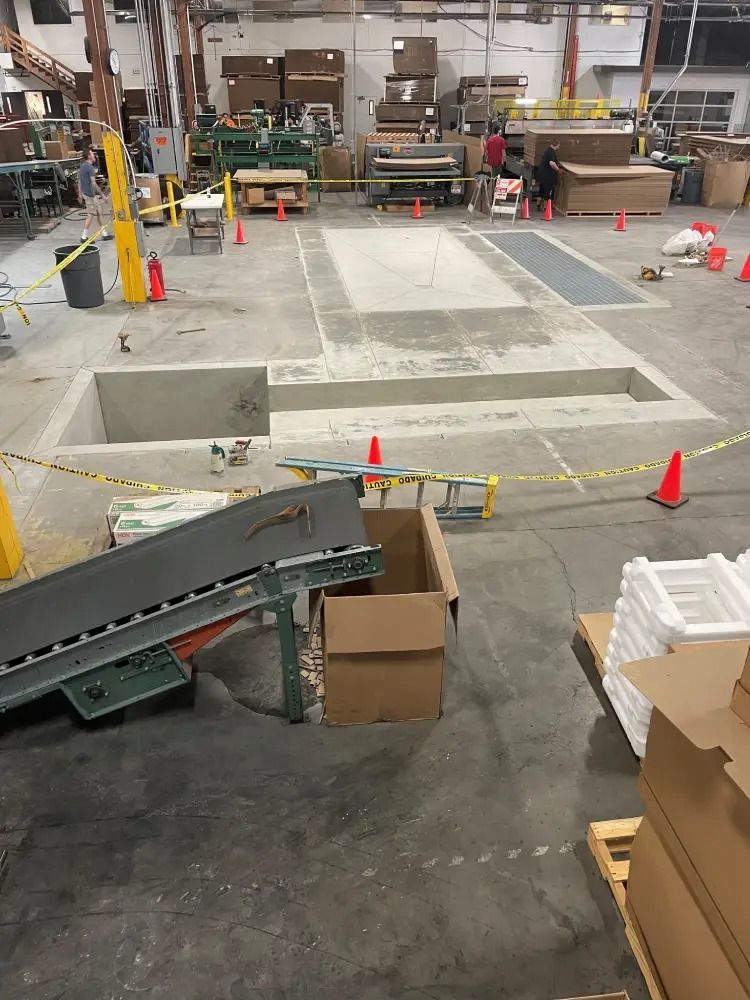 An industrial warehouse floor features a large, rectangular concrete cutout surrounded by orange safety cones and tape.