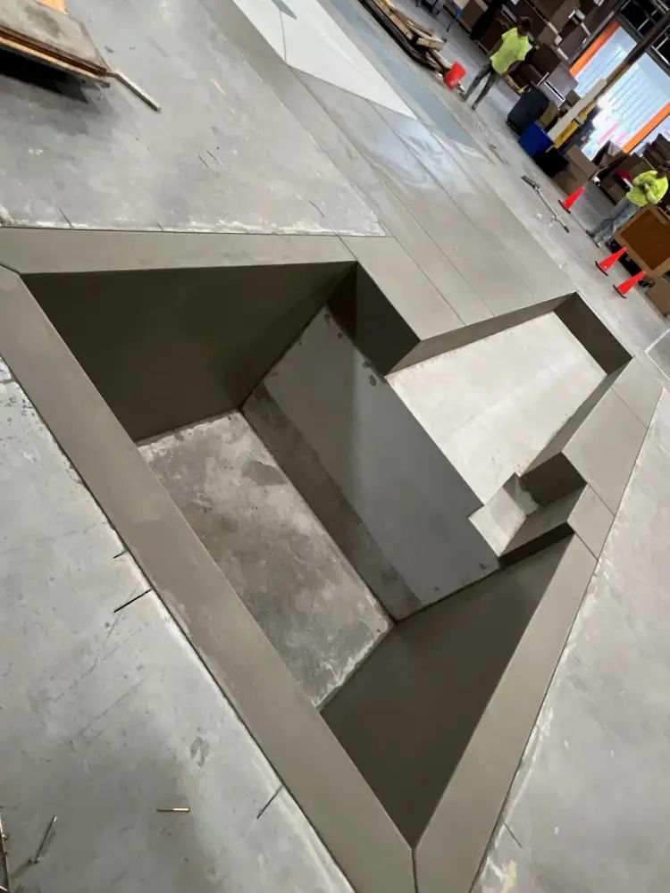 Concrete construction of an open stairwell or sunken area on a flat, industrial floor with workers in the background.