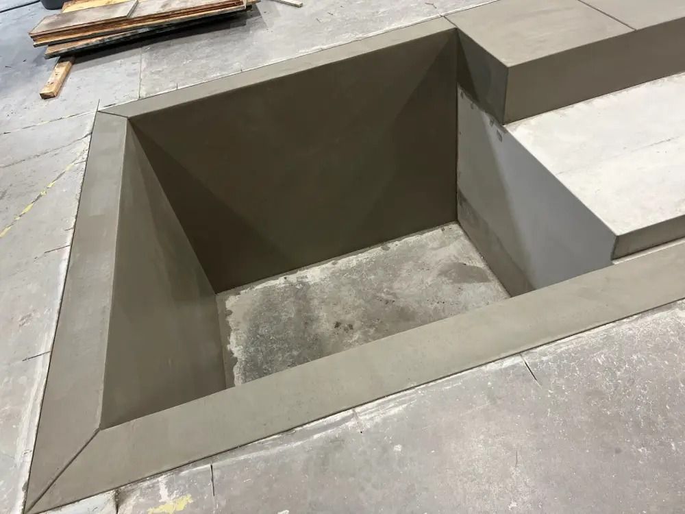 A square concrete pit recessed in a floor, featuring smooth, angled edges and a rough, dark interior base.