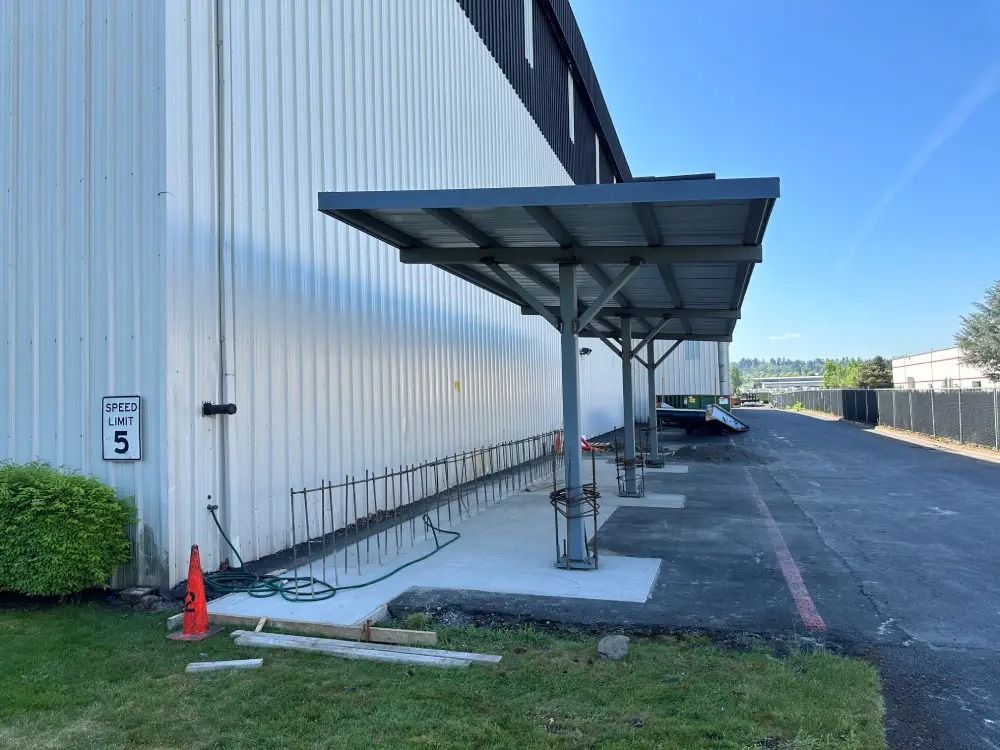 A covered bike rack installed on a concrete pad against the white metal wall of a building next to an asphalt road.